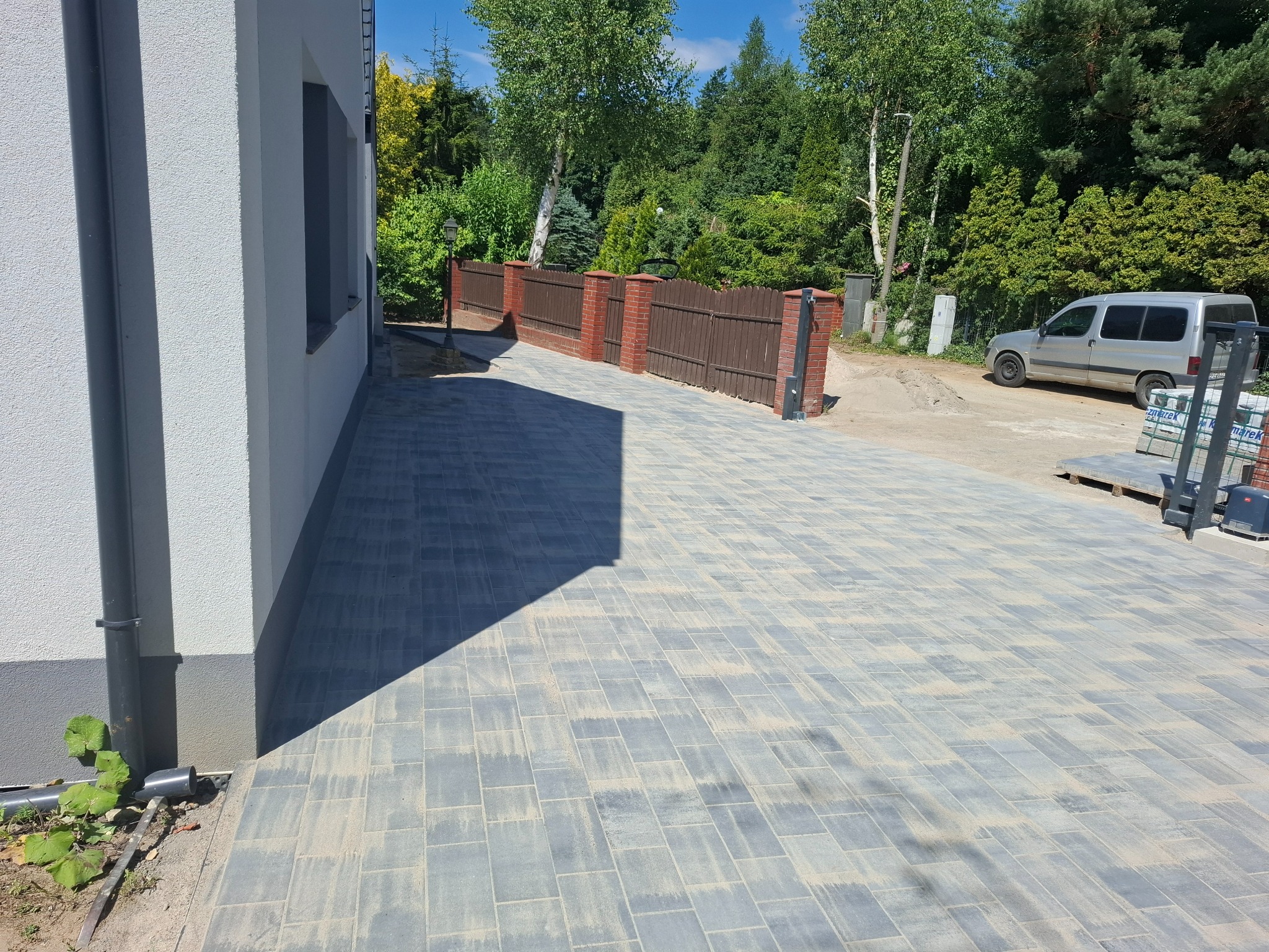 Ułożona szara kostka brukowa na podjeździe, widok z poziomu gruntu. W tle fragment budynku z rynną i ogrodzenie z ceglanymi słupkami. Samochód dostawczy w oddali.