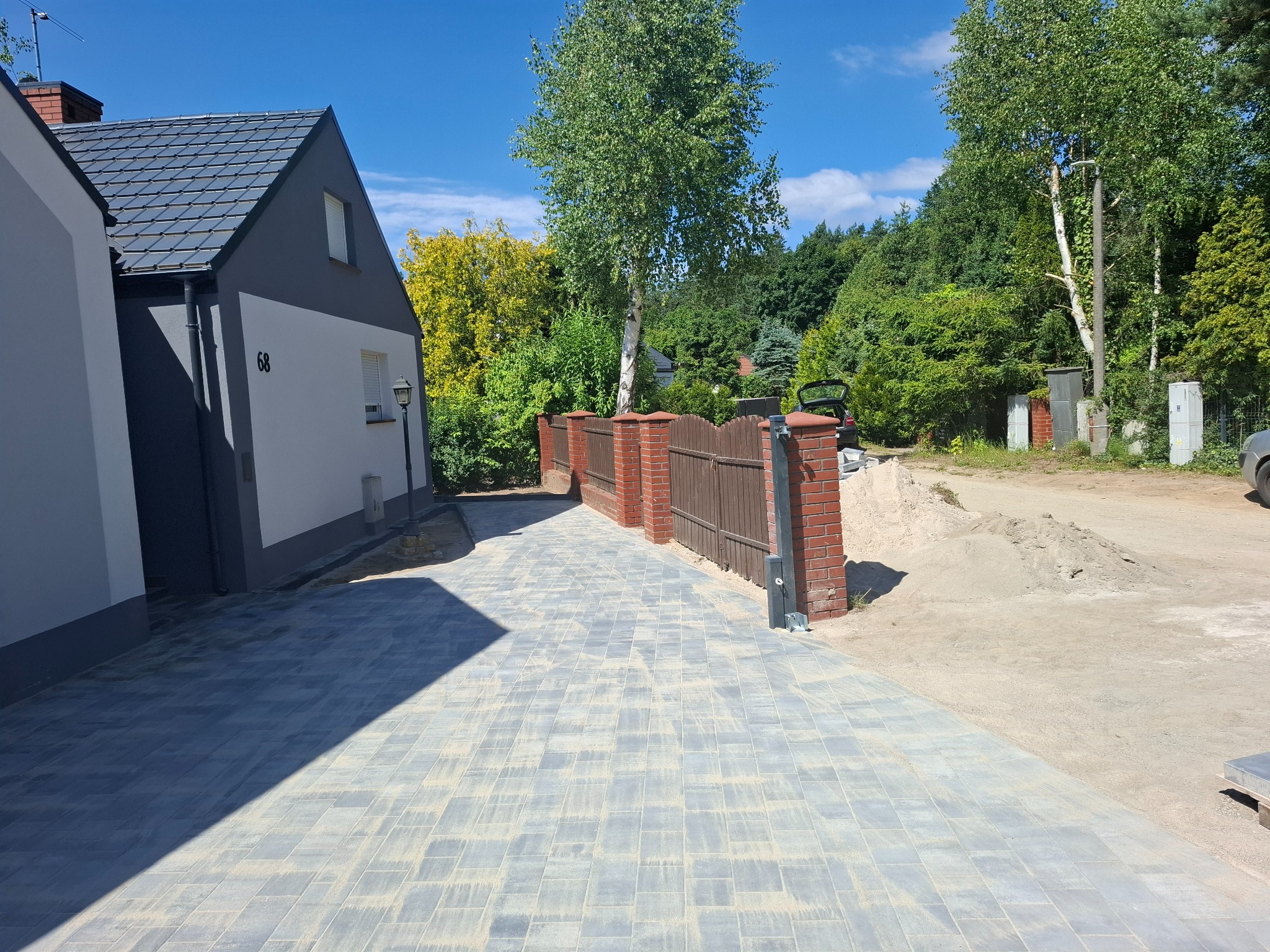 Ułożona szara kostka brukowa na podjeździe przed domem z numerem 68, z widocznym fragmentem ogrodzenia z cegły i drewnianą bramą. Słoneczny dzień.
