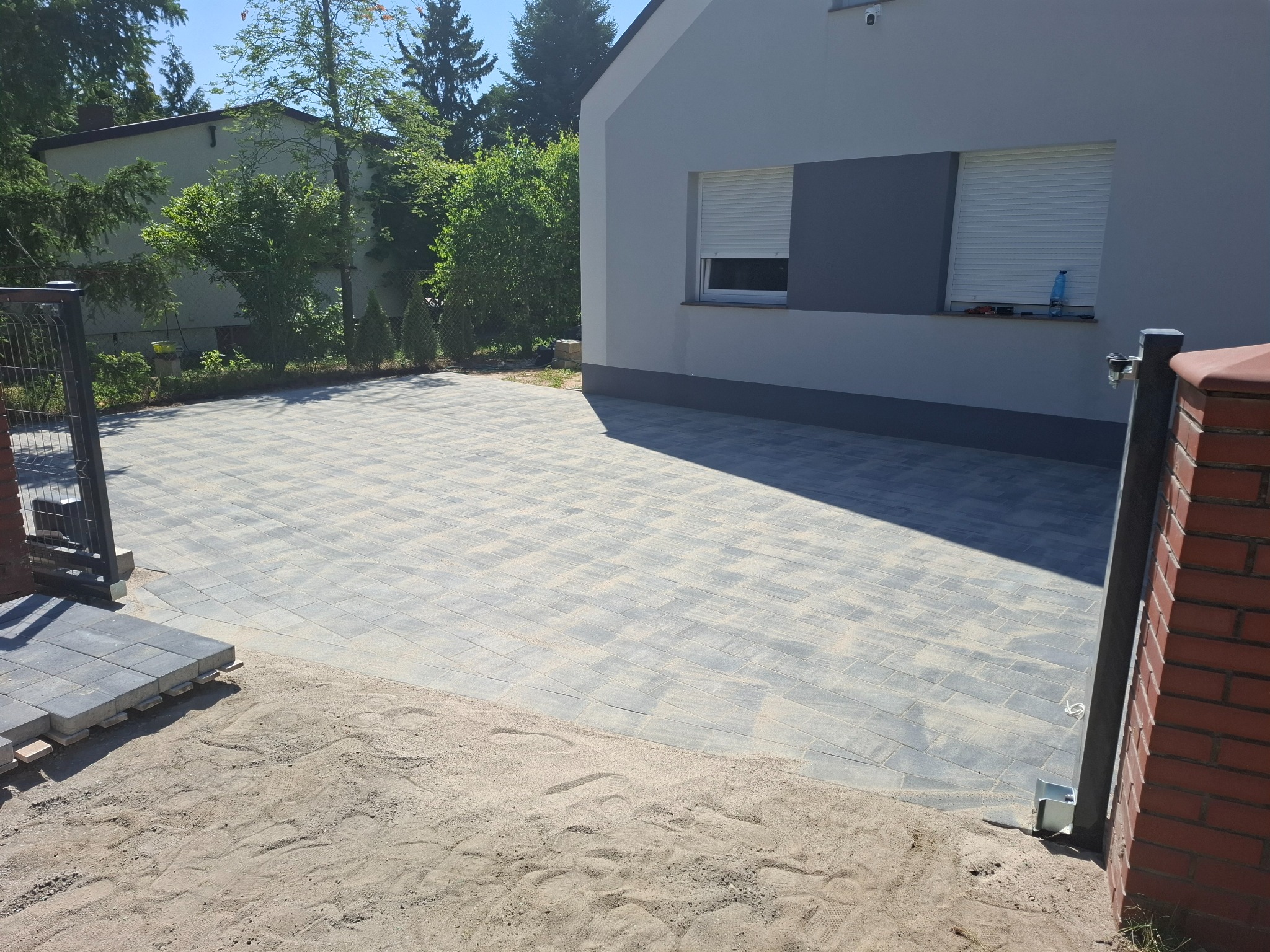 Ułożona szara kostka brukowa na podjeździe, z piaskiem na obrzeżach. W tle dom z szarą elewacją i zielenią. Widoczny fragment ogrodzenia i brama.