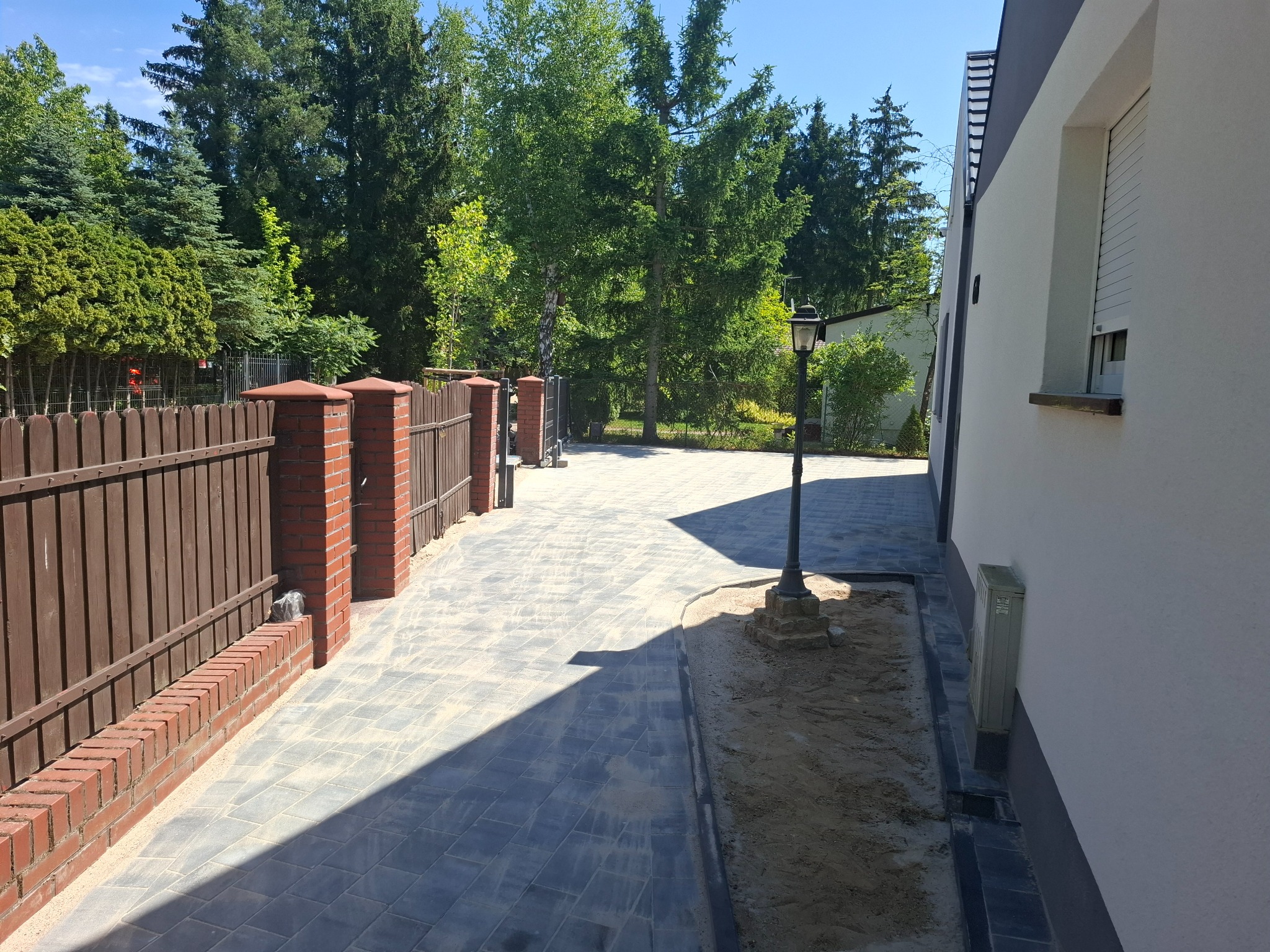 Ułożona kostka brukowa w szarym odcieniu tworzy ścieżkę wzdłuż budynku i ogrodzenia z ceglanymi słupkami. Widoczna lampa ogrodowa i zieleń drzew w tle.