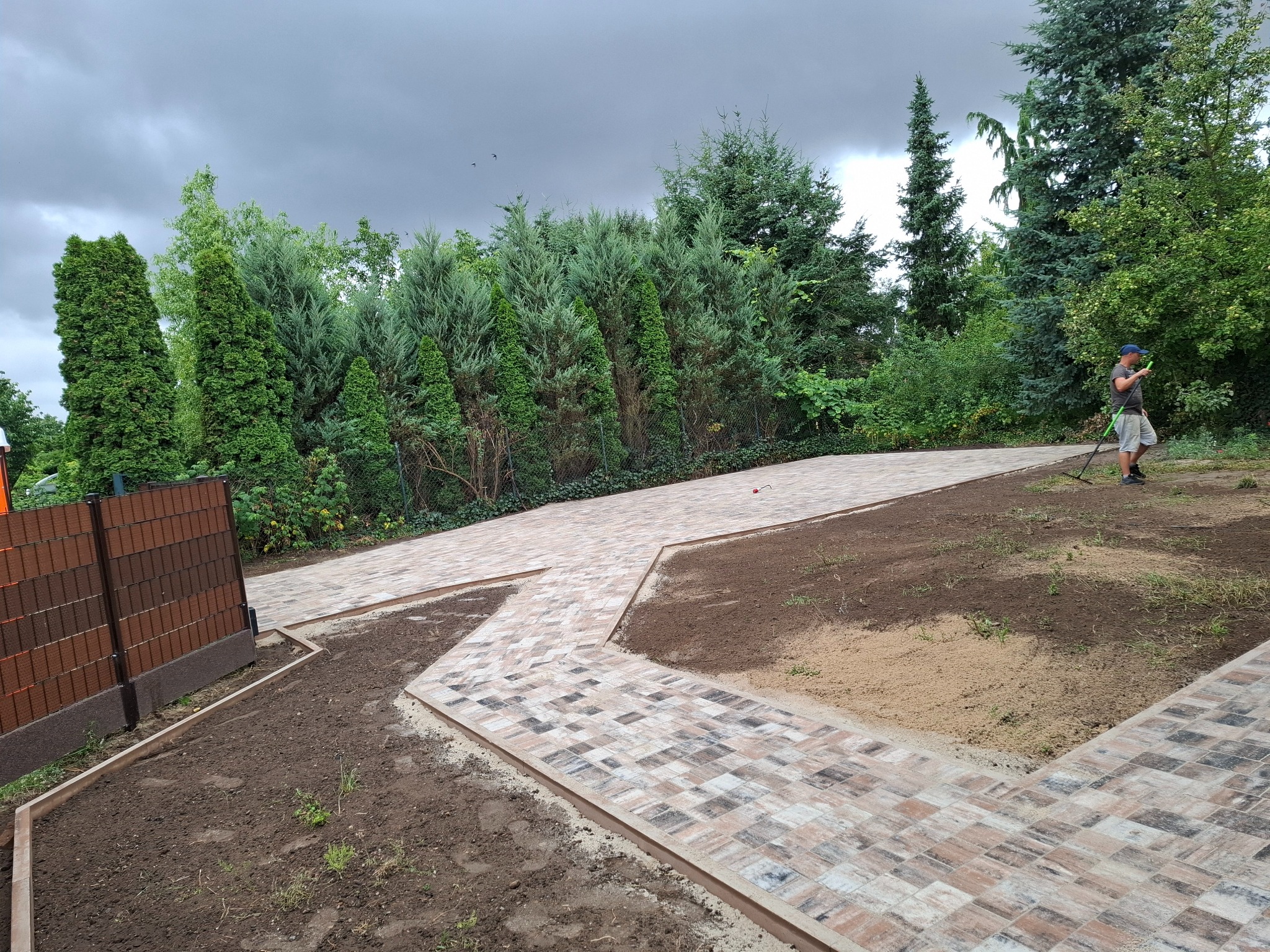 Kaskadowa ścieżka z kostki brukowej w ogrodzie, wykończona ozdobnymi obrzeżami. W tle zielone drzewa i krzewy, po prawej stronie osoba z narzędziem ogrodniczym.