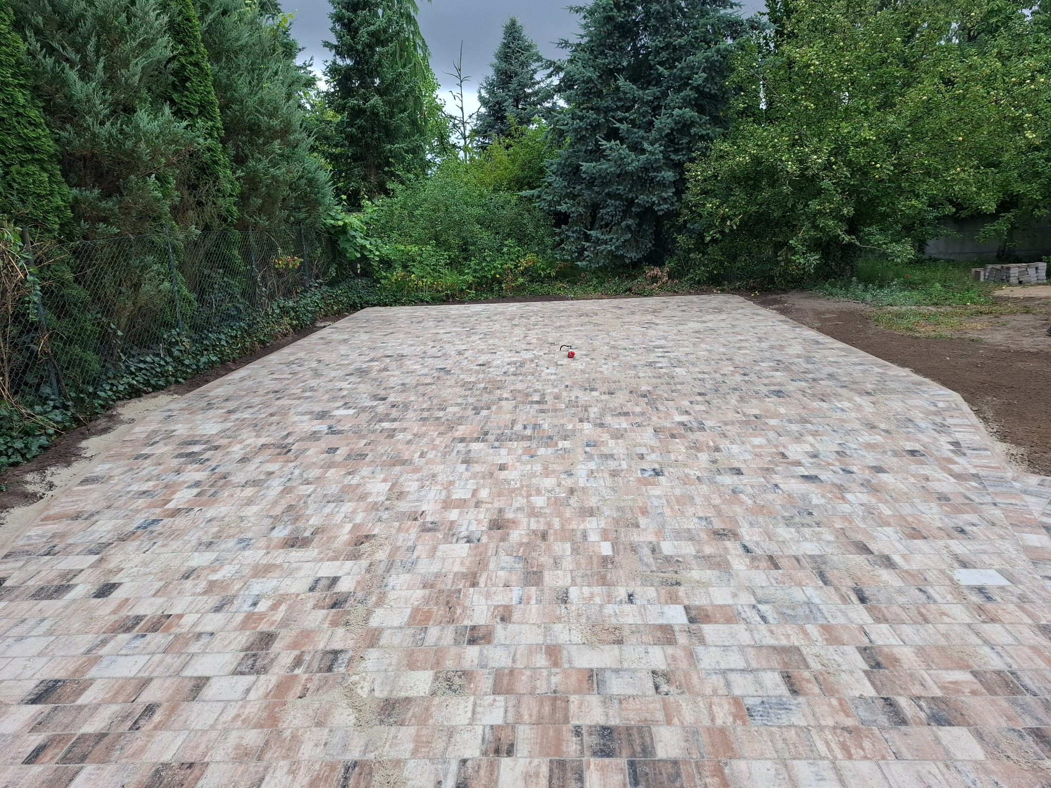 Nowo ułożona, wielobarwna kostka brukowa tworzy równą powierzchnię na tle zieleni drzew i krzewów. Widoczny czerwony element, prawdopodobnie znacznik.