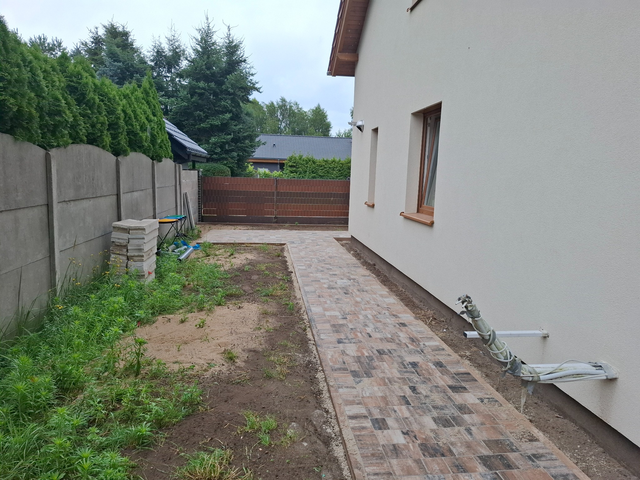 Ścieżka z kostki brukowej prowadząca od betonowego płotu do budynku z oknami. Stosy kostki, widoczne instalacje na ścianie.