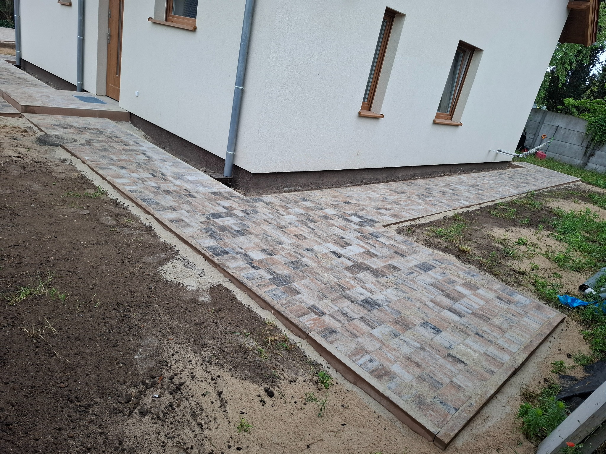 Ścieżka z kostki brukowej o nieregularnym wzorze prowadząca do domu z beżową elewacją i brązowymi oknami. Widoczne obrzeża i przygotowane podłoże.