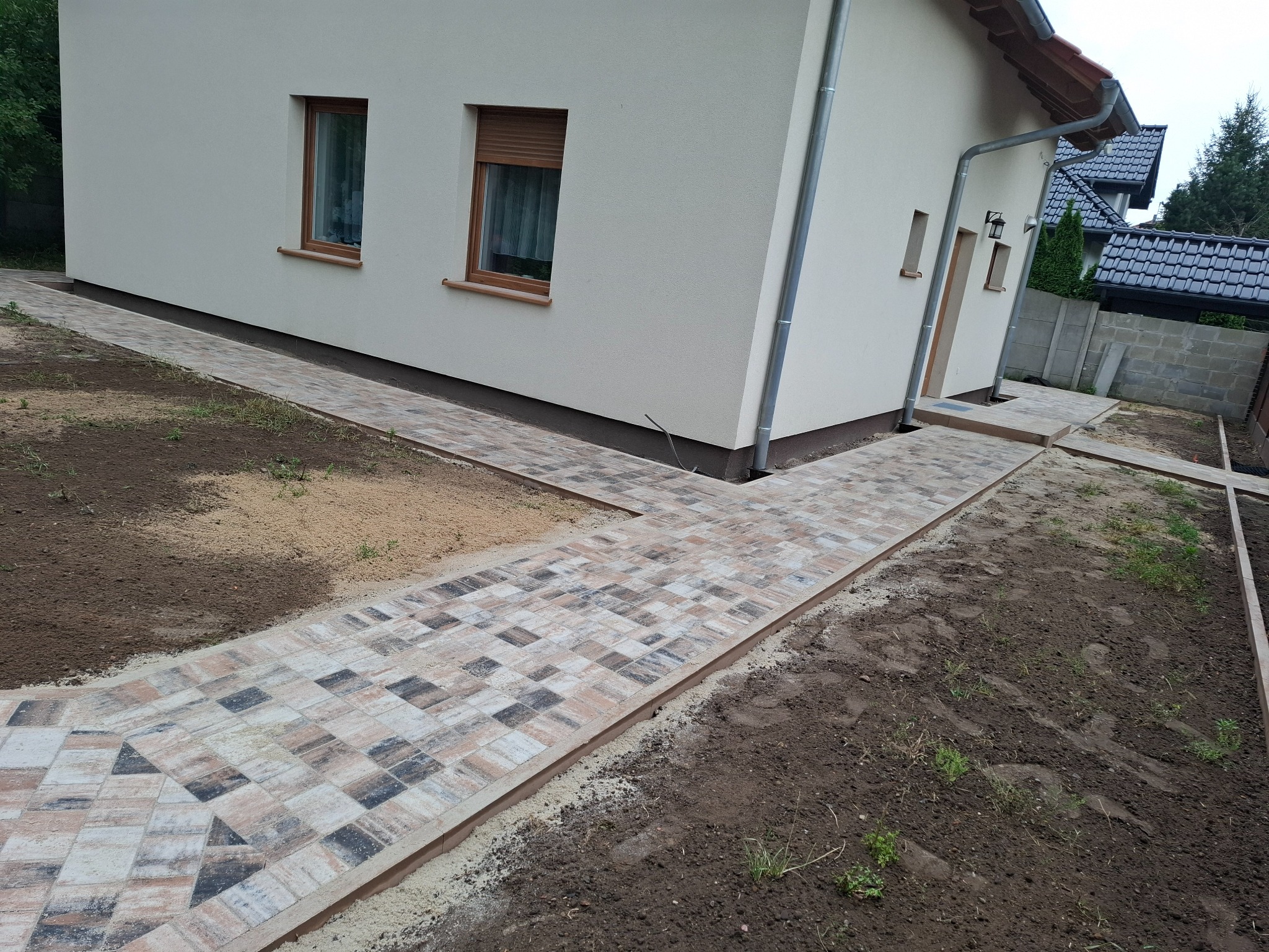 Ścieżka z kostki brukowej prowadząca do domu, z widocznymi detalami ułożenia i estetycznym wykończeniem obrzeży. Tło stanowi elewacja budynku i przygotowany teren.