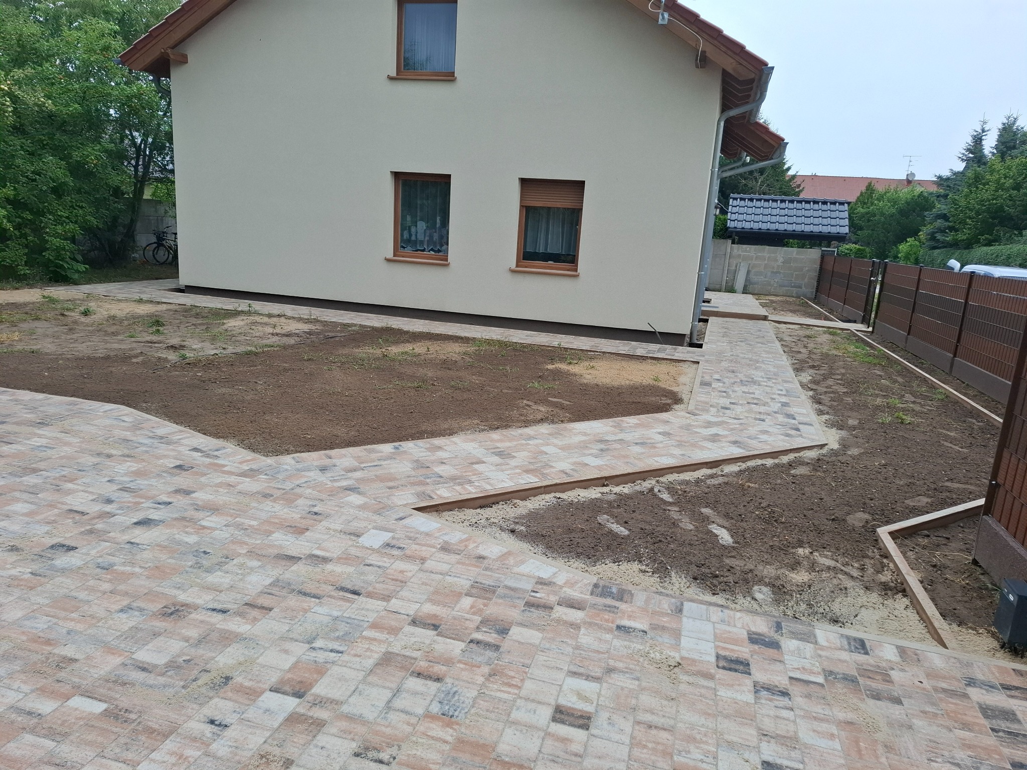 Ścieżka z kostki brukowej o nieregularnym wzorze prowadząca wokół domu z beżową elewacją i brązowymi oknami, widok z perspektywy ziemi, częściowo wykończona.