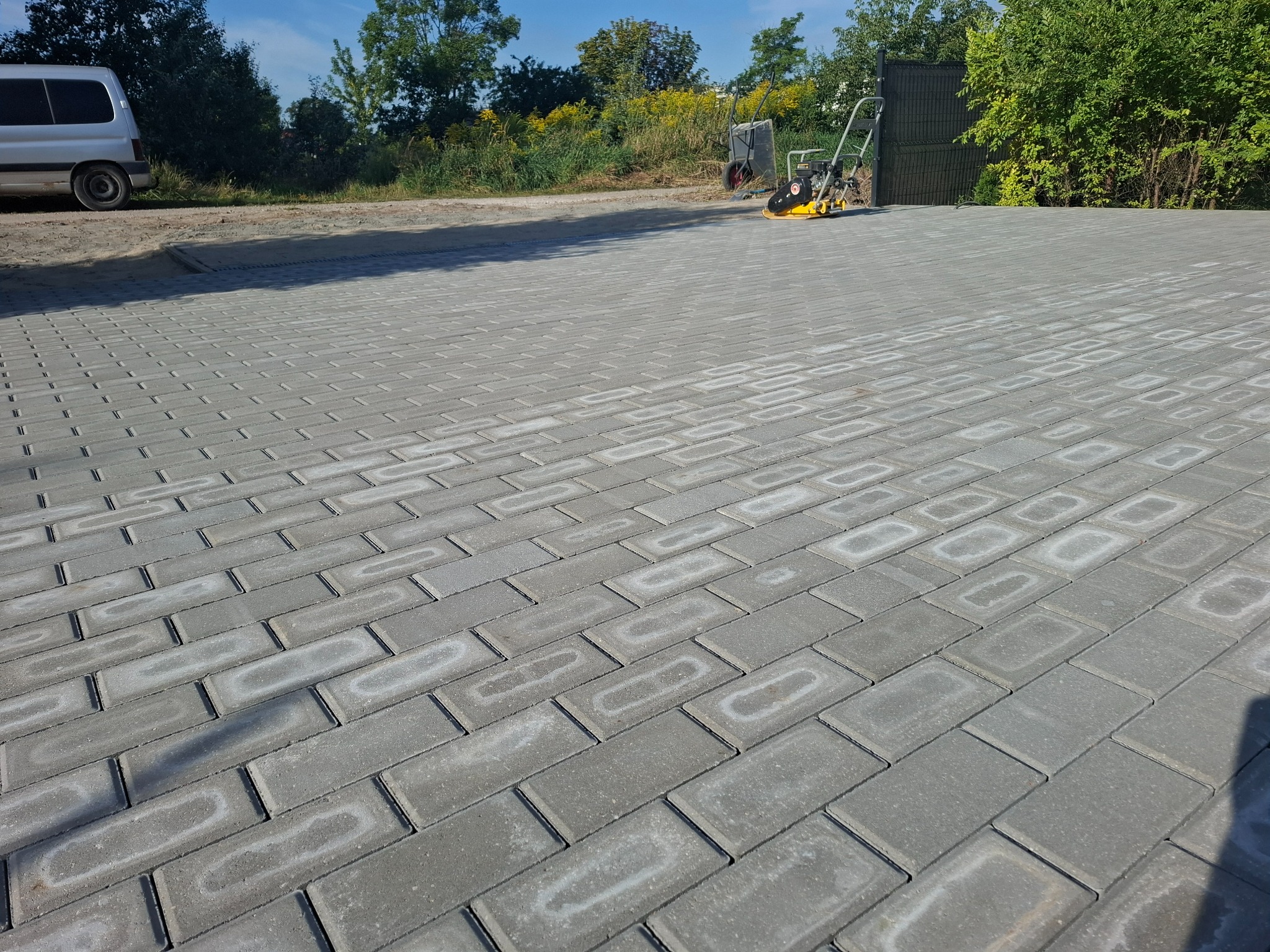 Ułożona szara kostka brukowa na posesji, z widoczną ubijarką w tle, sugerującą etap prac wykończeniowych. W tle zieleń i częściowo widoczny biały samochód.