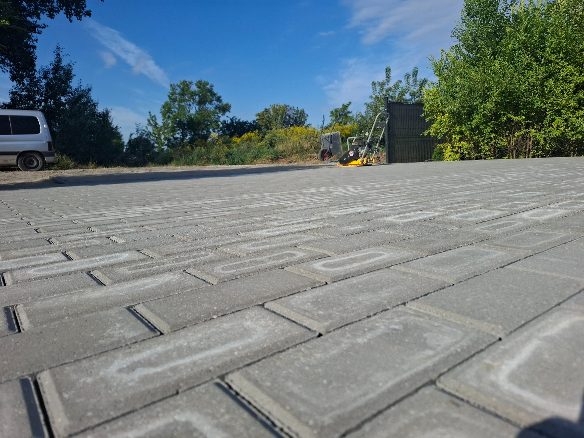Nowa, szara kostka brukowa ułożona w regularny wzór. W tle widać zagęszczarkę, wózek i samochód dostawczy na tle zieleni i błękitnego nieba.