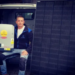 Mężczyzna siedzi w bagażniku białego samochodu dostawczego, trzymając falownik Fronius Symo z naklejką Solaris Fotowoltaika i stojąc obok panelu słonecznego.