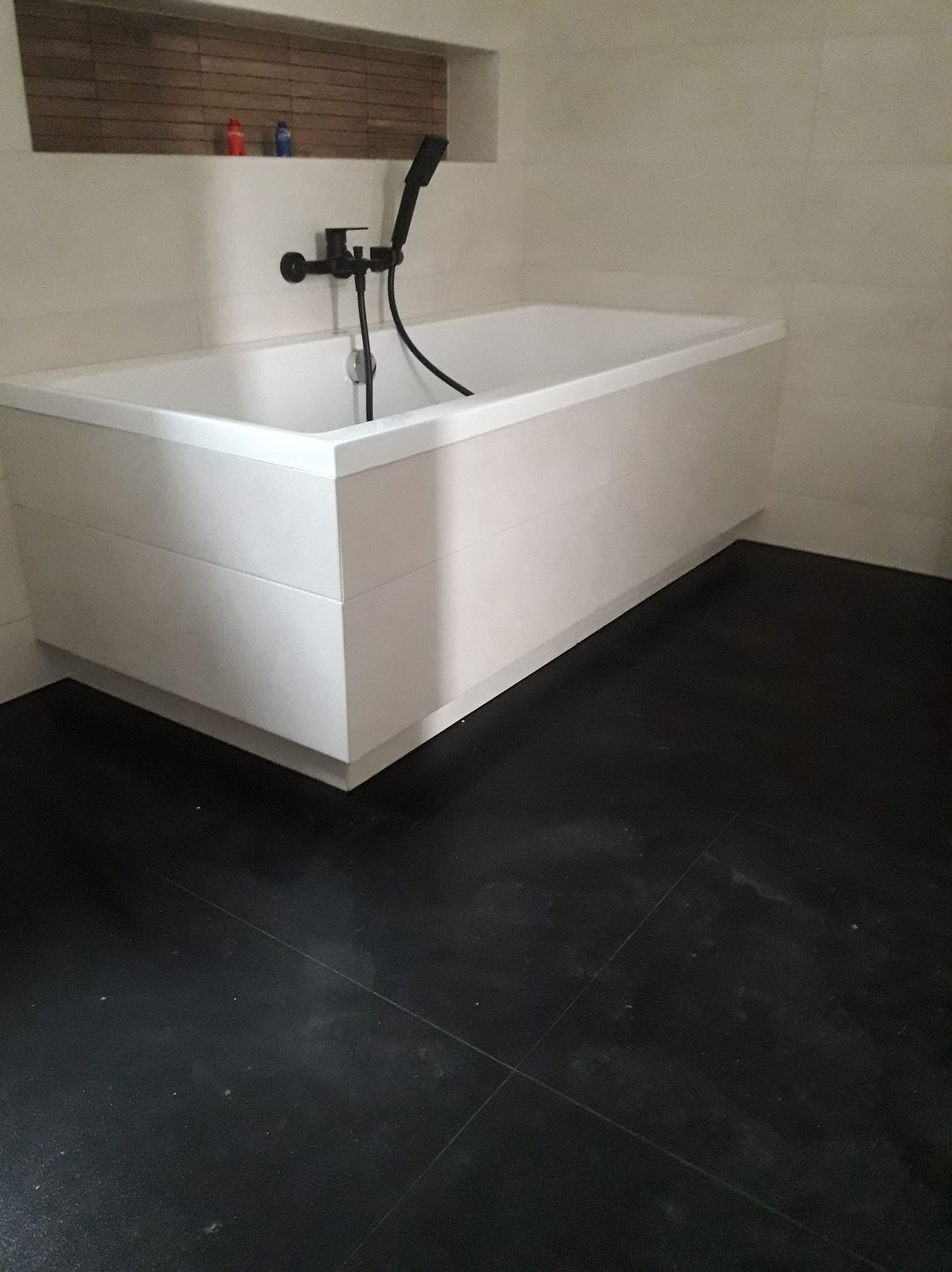 Nowoczesna, prostokątna wanna obudowana białymi panelami, z czarną armaturą i prysznicem, na tle jasnych płytek i ciemnej podłogi z płytek. W tle widoczna półka z drewnianym wykończeniem i dwoma...
