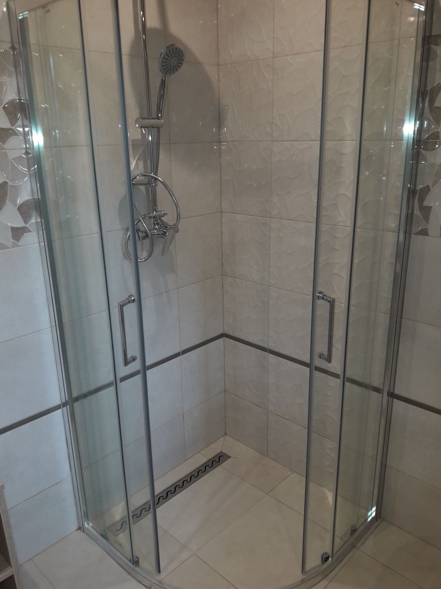 Narożna kabina prysznicowa z chromowaną armaturą i odpływem liniowym w posadzce wyłożonej beżowymi płytkami z delikatnym wzorem.