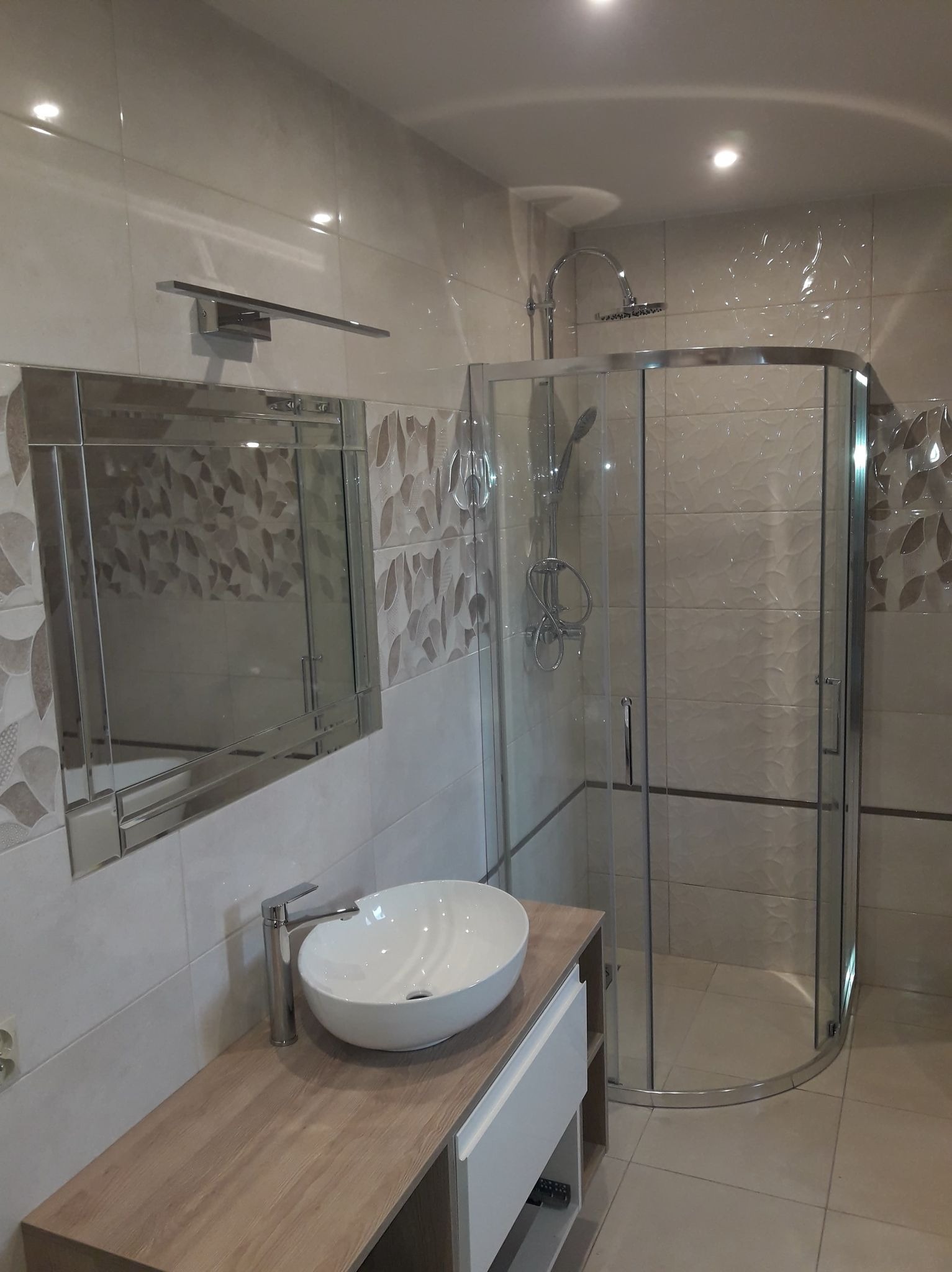 Nowoczesna łazienka z kabiną prysznicową z zaokrąglonymi szybami, umywalką nablatową na drewnianym blacie i lustrem z oświetleniem, wykończona płytkami w beżowych odcieniach z dekoracyjnym wzorem...