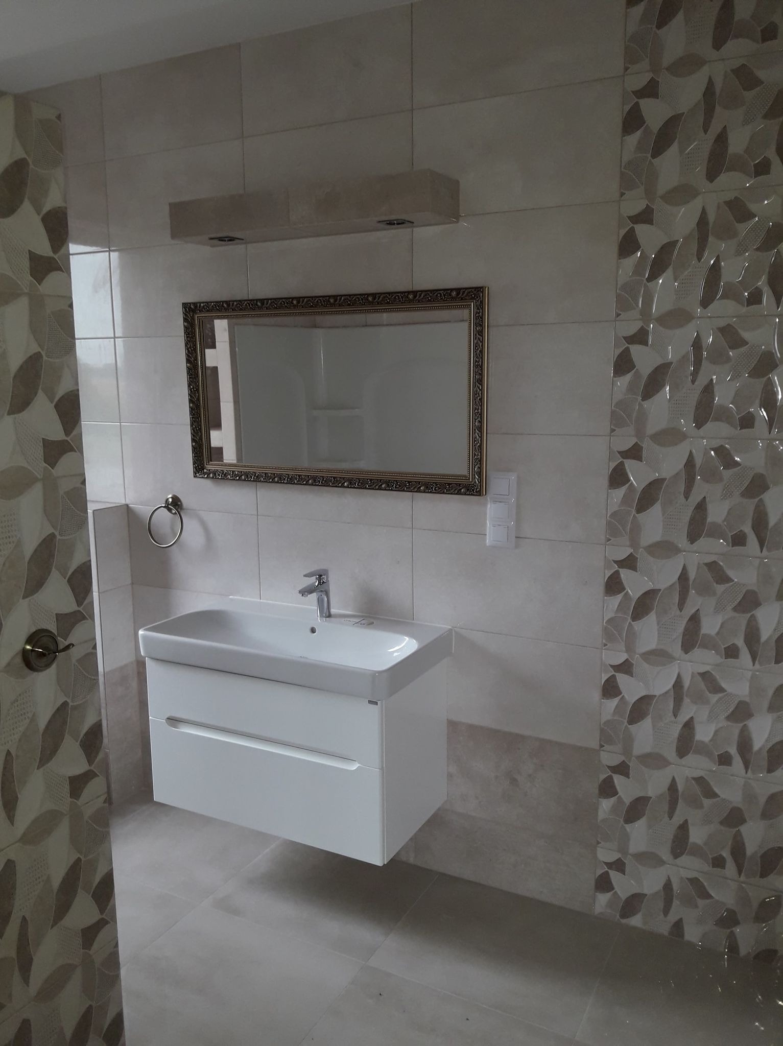 Nowoczesna łazienka z białym blatem umywalki, lustrem w złotej ramie i dekoracyjnymi płytkami imitującymi liście na ścianie.