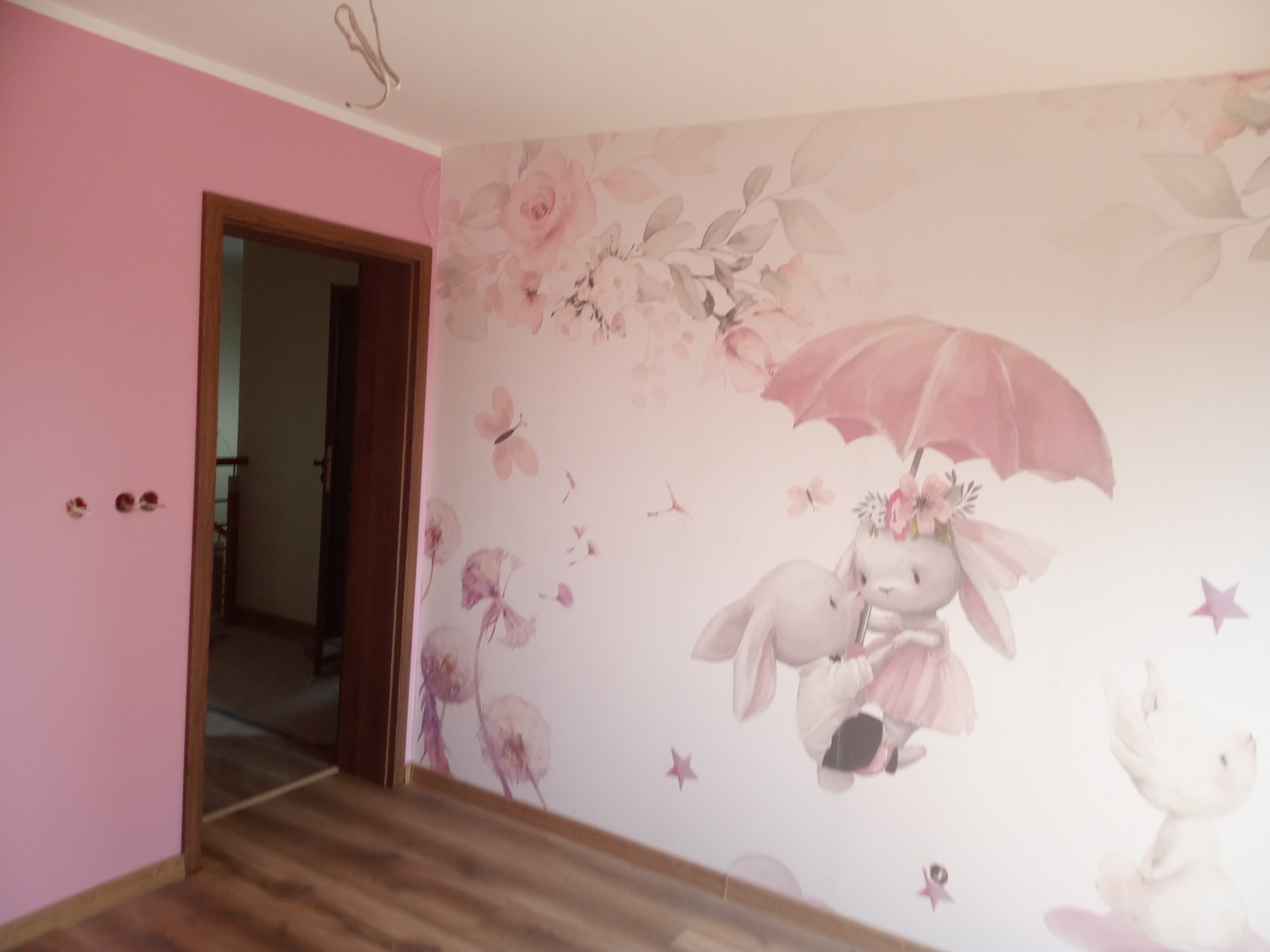 Pokój dziecięcy po odświeżeniu: Różowa ściana z trzema otworami po gniazdkach, drewniana podłoga i fototapeta z motywem królików pod parasolem.