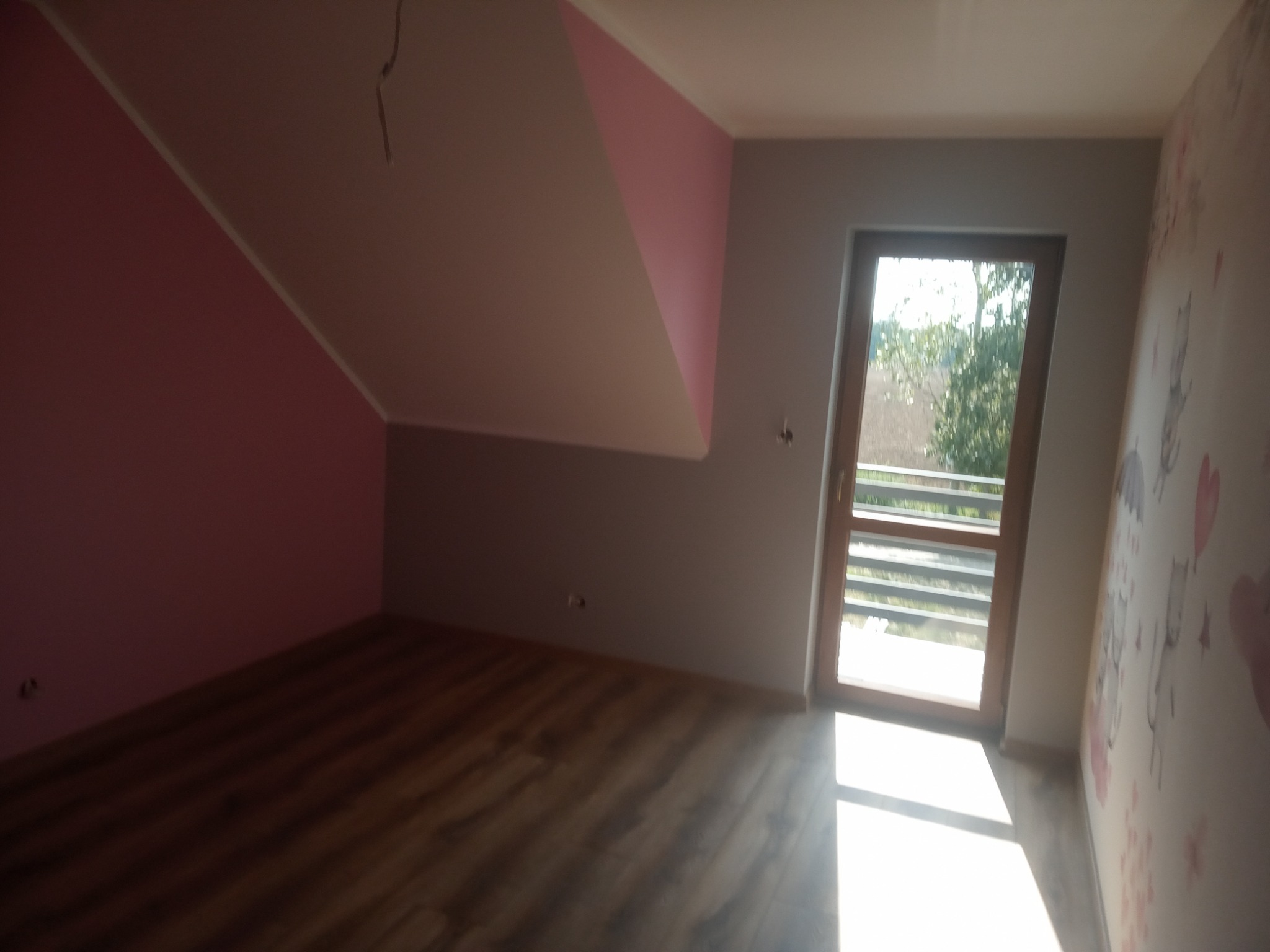 Pomalowane na różowo i szaro poddasze ze skośnym sufitem, widoczne panele podłogowe i okno balkonowe.