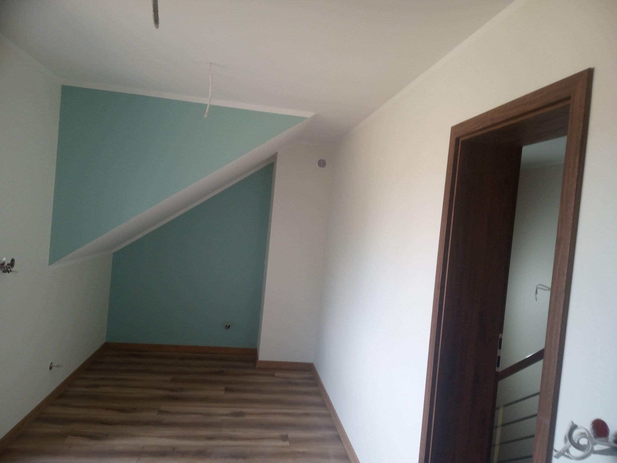Pokój na poddaszu po remoncie: świeżo pomalowana ściana w kolorze turkusowym, ułożone panele podłogowe w odcieniach brązu, widoczny fragment drewnianej futryny drzwiowej.