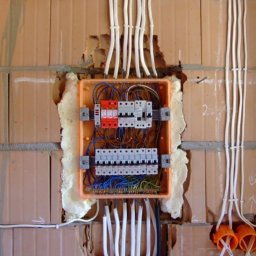 Oleg Vahniuk - Pomarańczowa skrzynka rozdzielcza z zabezpieczeniami i przewodami zamontowana na ścianie z cegły, zabezpieczona pianką montażową, z widocznymi przewodami w peszlach mocowanych opaskami.