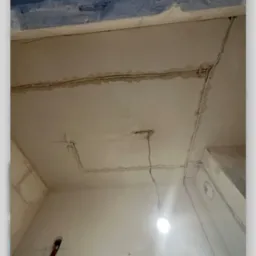 Instalacja elektryczna w trakcie montażu w surowym wnętrzu, widoczne kable prowadzone po suficie i ścianach, częściowo zamaskowane gipsem, z wiszącą żarówką.