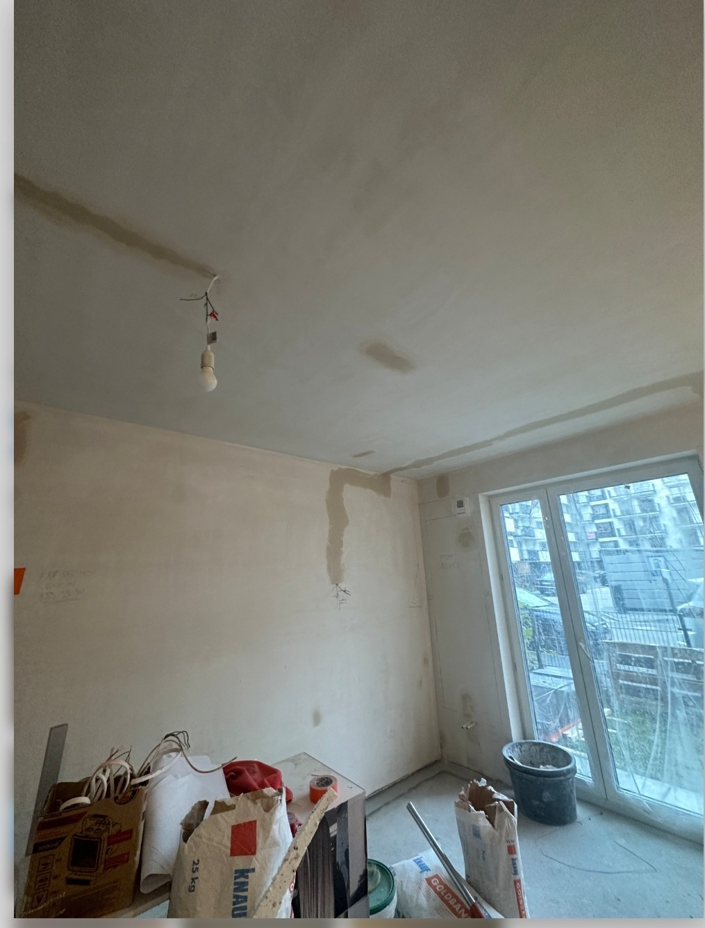 Surowe wnętrze w trakcie remontu: widoczne worki z zaprawą Knauf i Goldsan, prowizoryczna żarówka zwisająca z sufitu, ślady po instalacjach elektrycznych na ścianie, okno z widokiem na budynki.