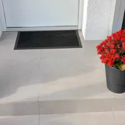 Jasne płytki ceramiczne na schodach wejściowych z czarną wycieraczką, doniczkami z czerwonymi kwiatami i dynią po lewej stronie.