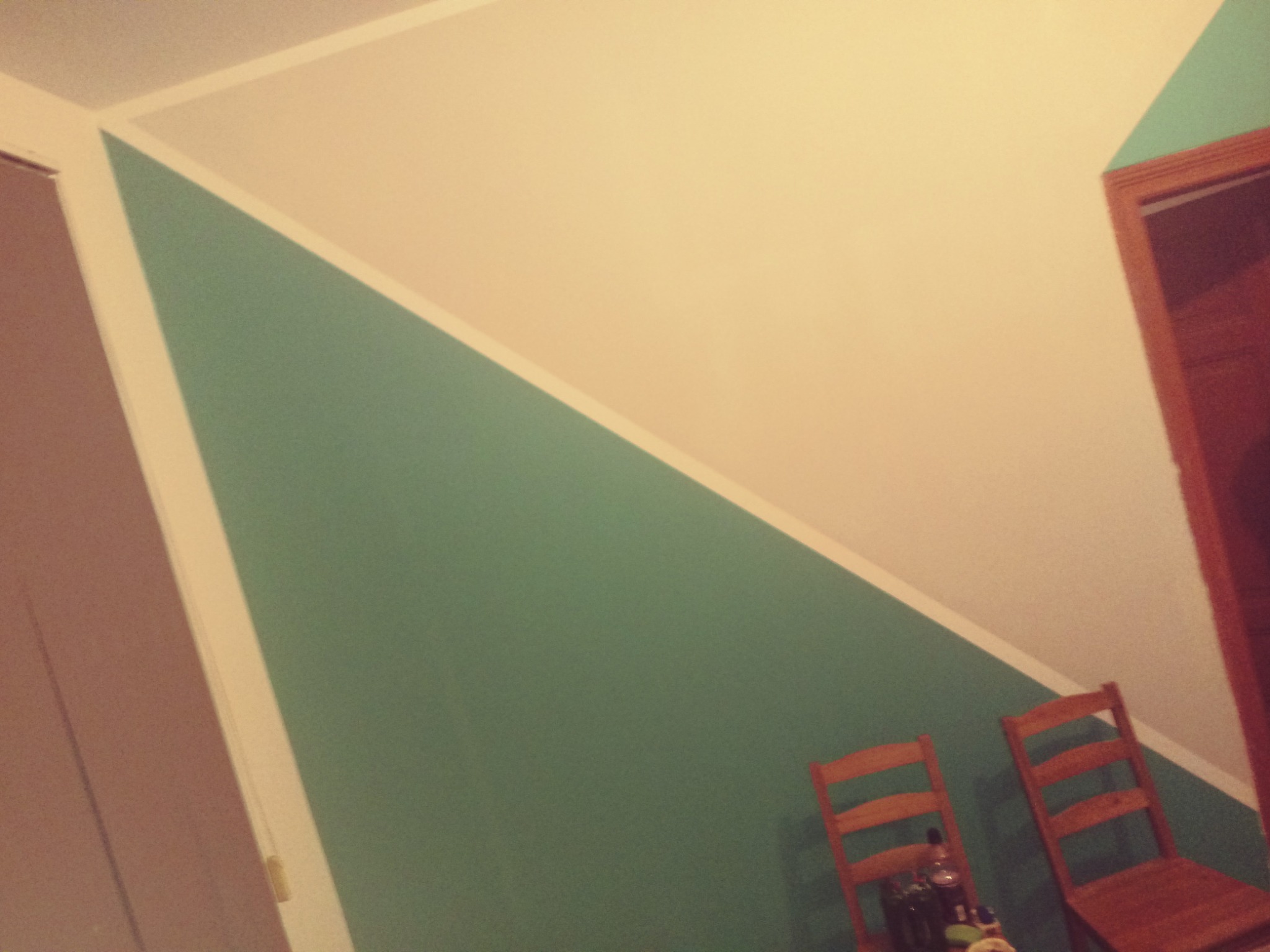 Ściana pomalowana na dwa kolory, zielony i kremowy, z ukośnym podziałem. Widoczne dwie drewniane krzesła przy ścianie i fragment ościeżnicy drzwi.