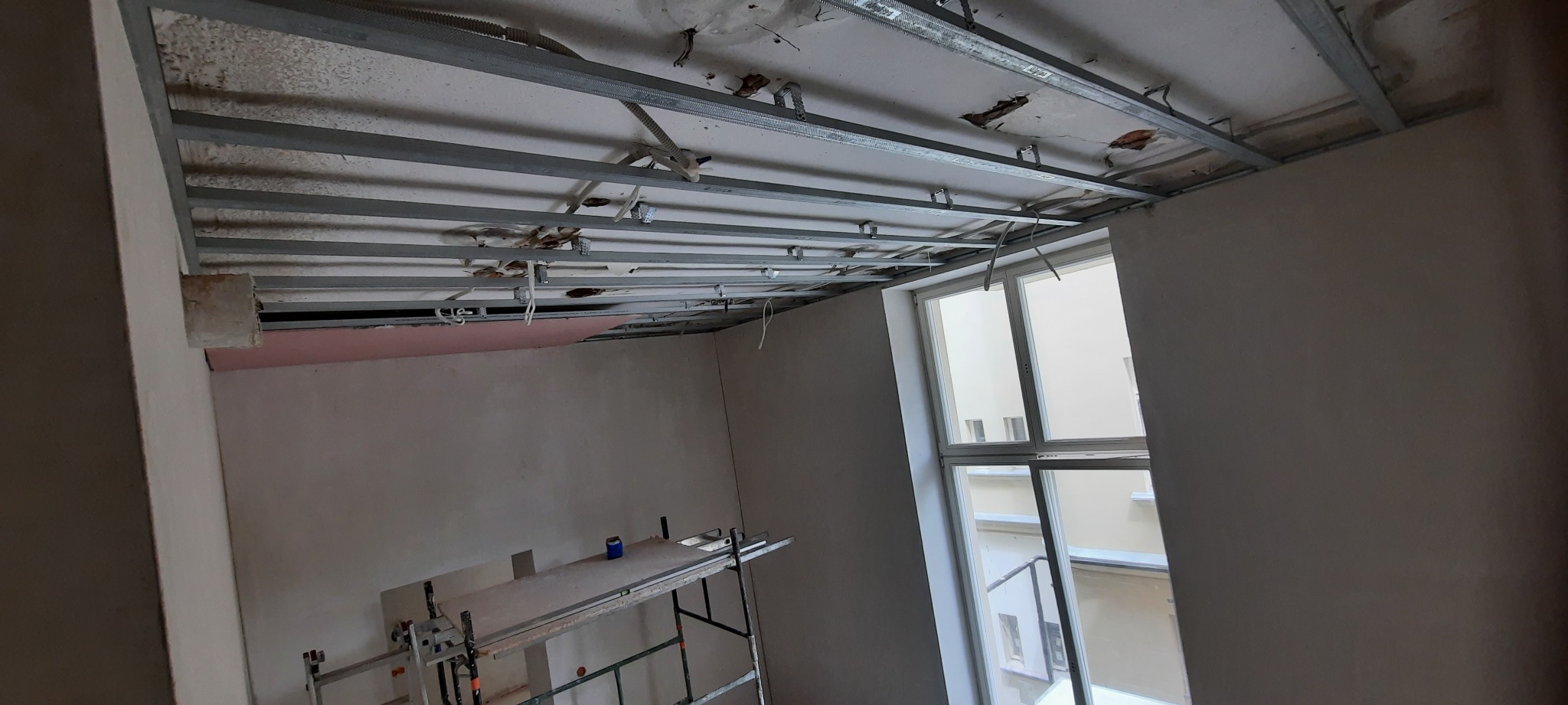 Stelaż sufitu podwieszanego z widocznymi fragmentami starego sufitu, częściowo zakrytego płytą, oraz rusztowanie w pomieszczeniu z oknem.