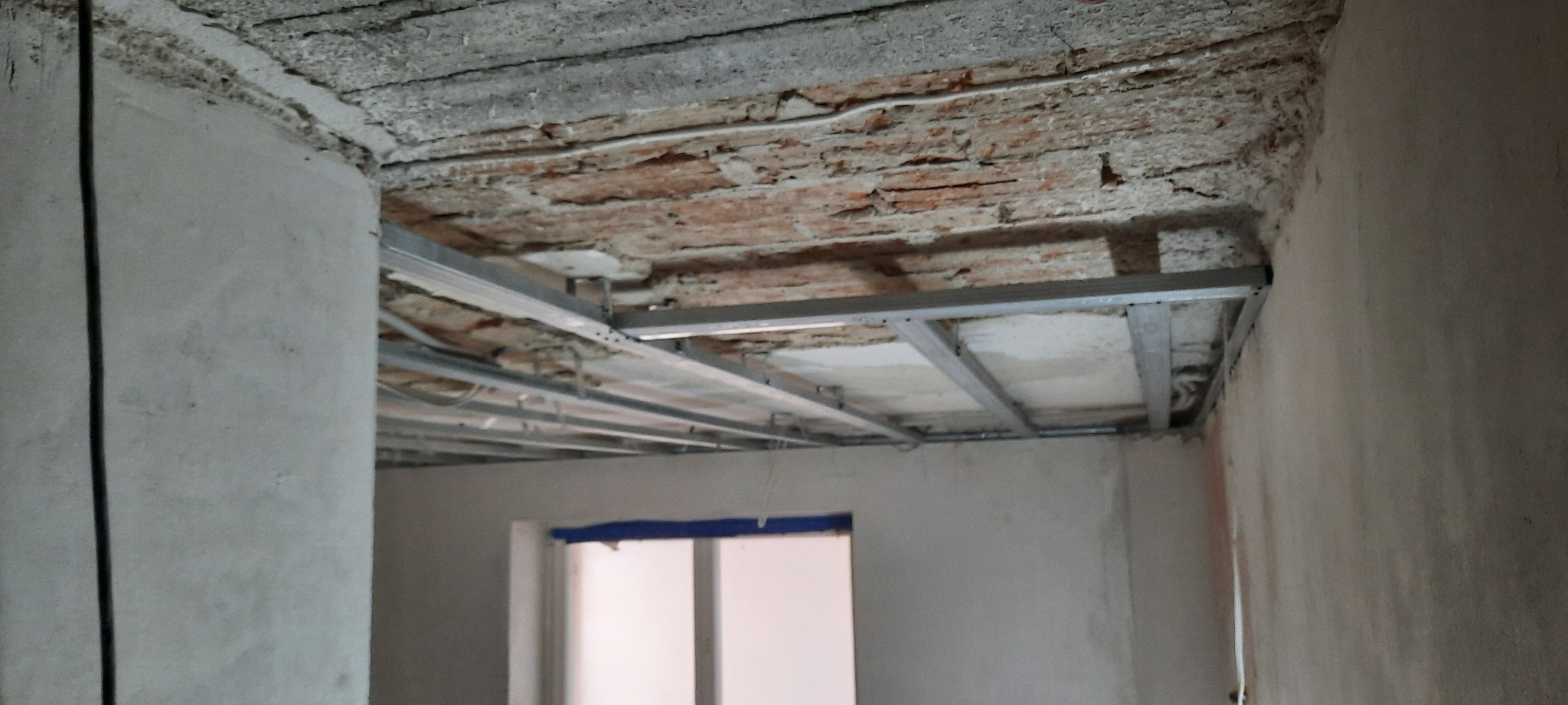 Niedokończony sufit z widoczną konstrukcją metalową i fragmentami starego, ceglanego stropu w remontowanym pomieszczeniu z oknem.