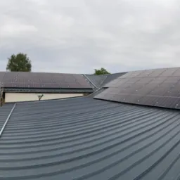 Panele słoneczne zamontowane na falistym, metalowym dachu budynku gospodarczego, widoczne rusztowanie i silos.