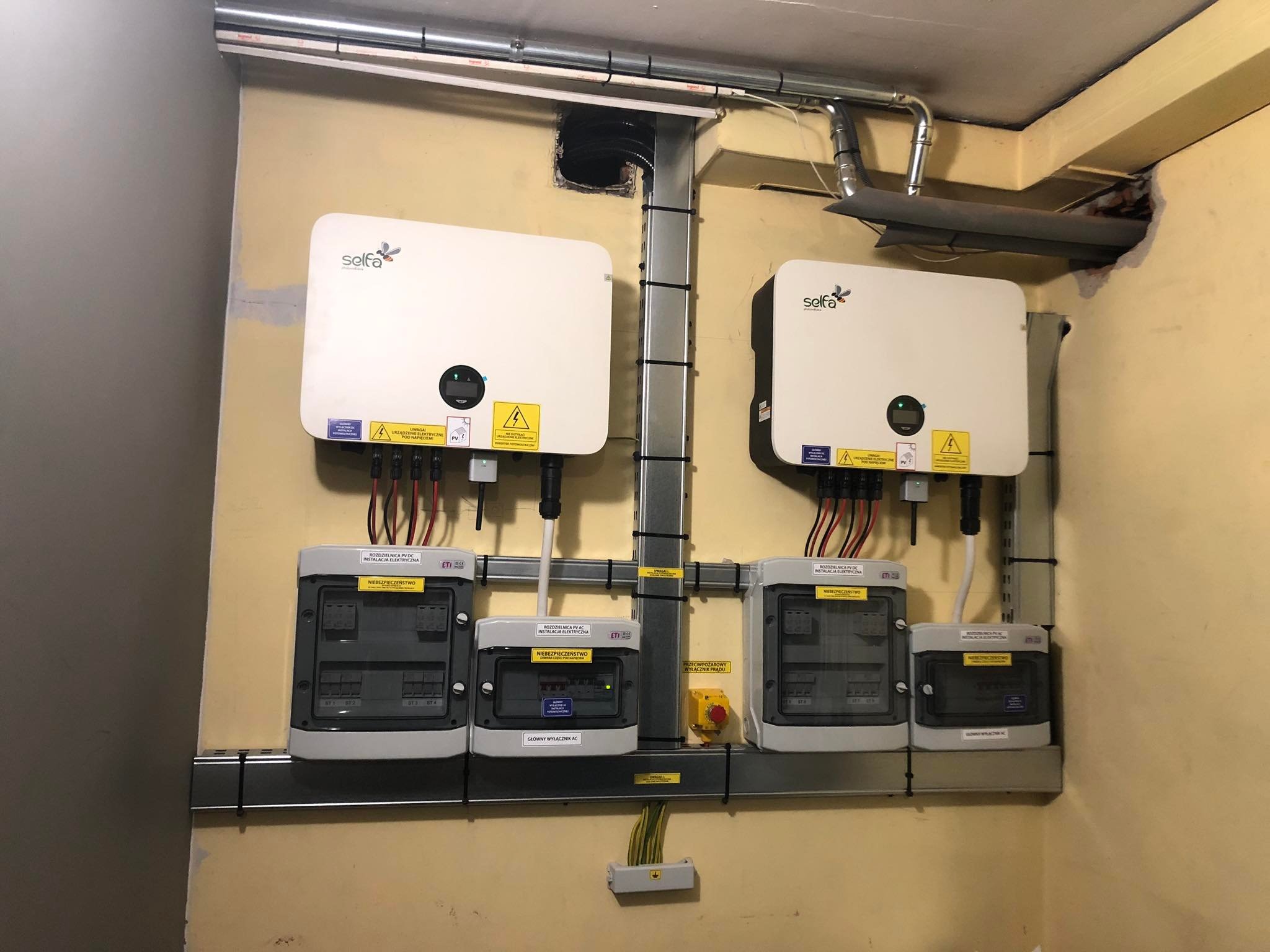 Dwa inwertery z logo firmy i rozdzielnice prądu stałego zamontowane na ścianie w pomieszczeniu technicznym, widoczne kable i oznaczenia ostrzegawcze.