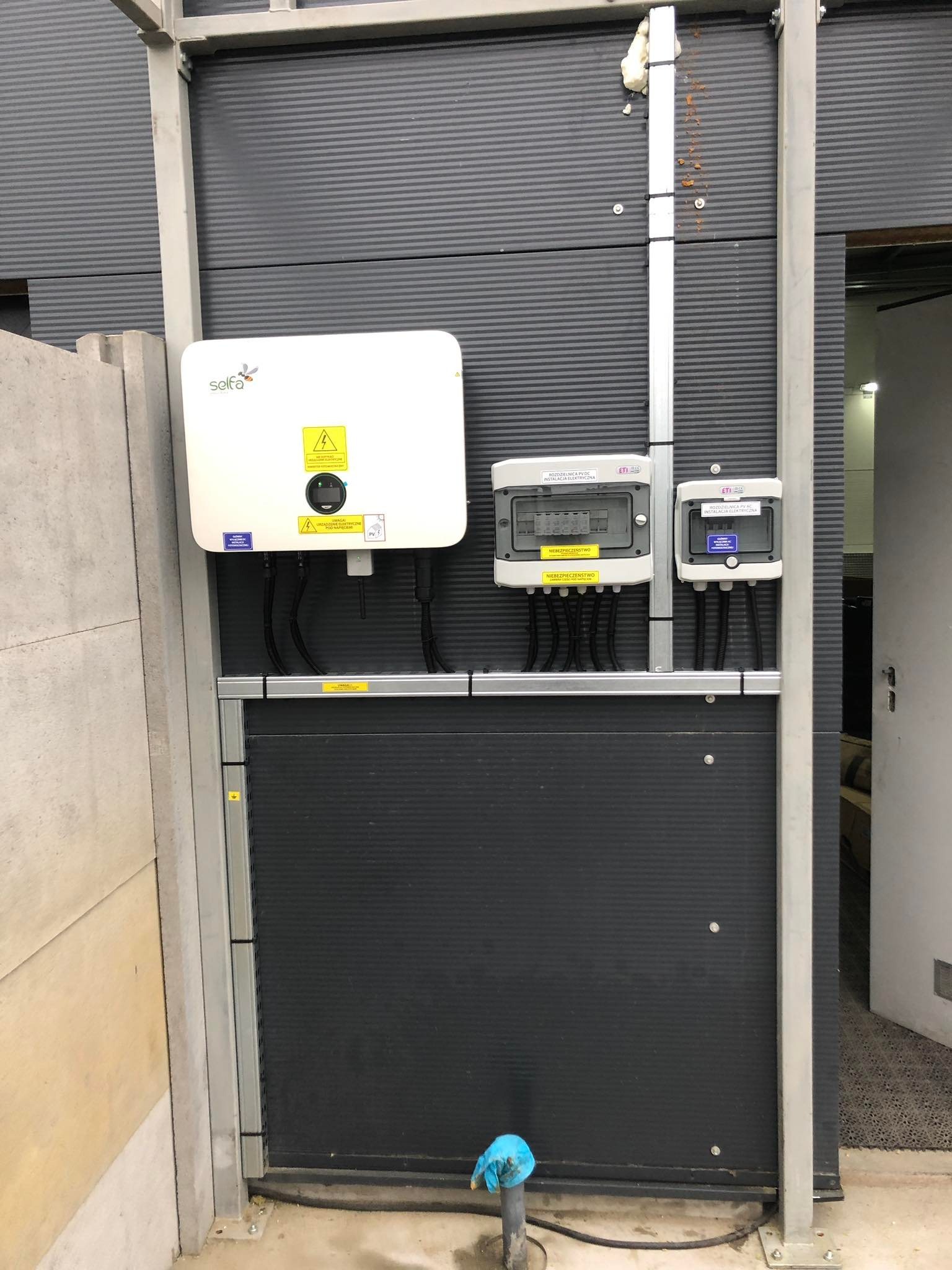 Zamontowany inwerter fotowoltaiczny marki Solfa, rozdzielnica PV DC i rozdzielnica AC na ciemnoszarej, ryflowanej ścianie w pomieszczeniu technicznym.