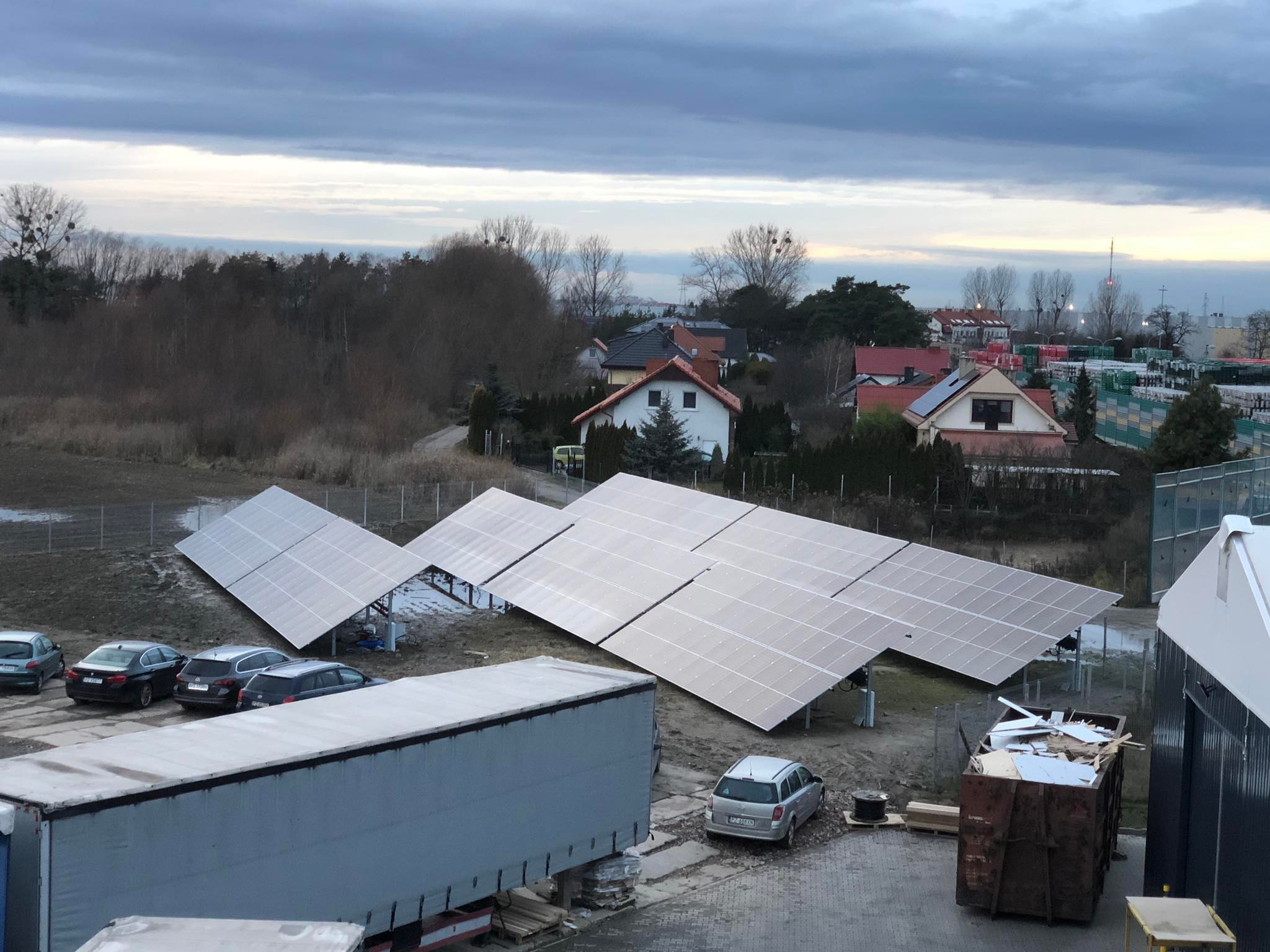 Rząd paneli słonecznych zamontowanych na gruncie, z widocznymi samochodami, przyczepą i kontenerem na odpady, na tle zabudowań mieszkalnych i drzew pod pochmurnym niebem.