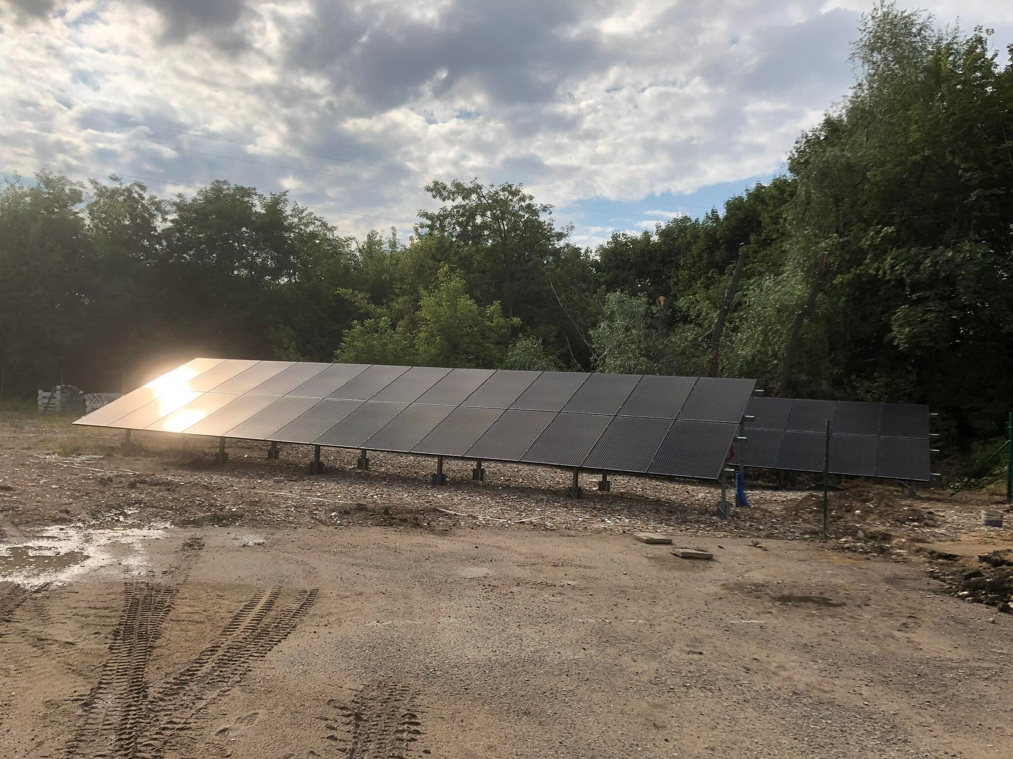 Panel słoneczny na metalowej konstrukcji, odbijający słońce, na tle drzew i pochmurnego nieba, widoczne ślady opon na ziemi.
