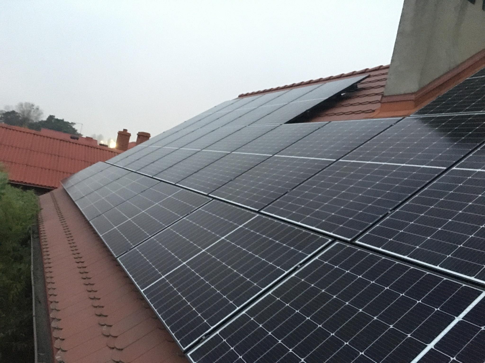 Panele słoneczne na czerwonym dachu domu jednorodzinnego, widoczne kominy i drzewa w tle, pochmurne niebo.