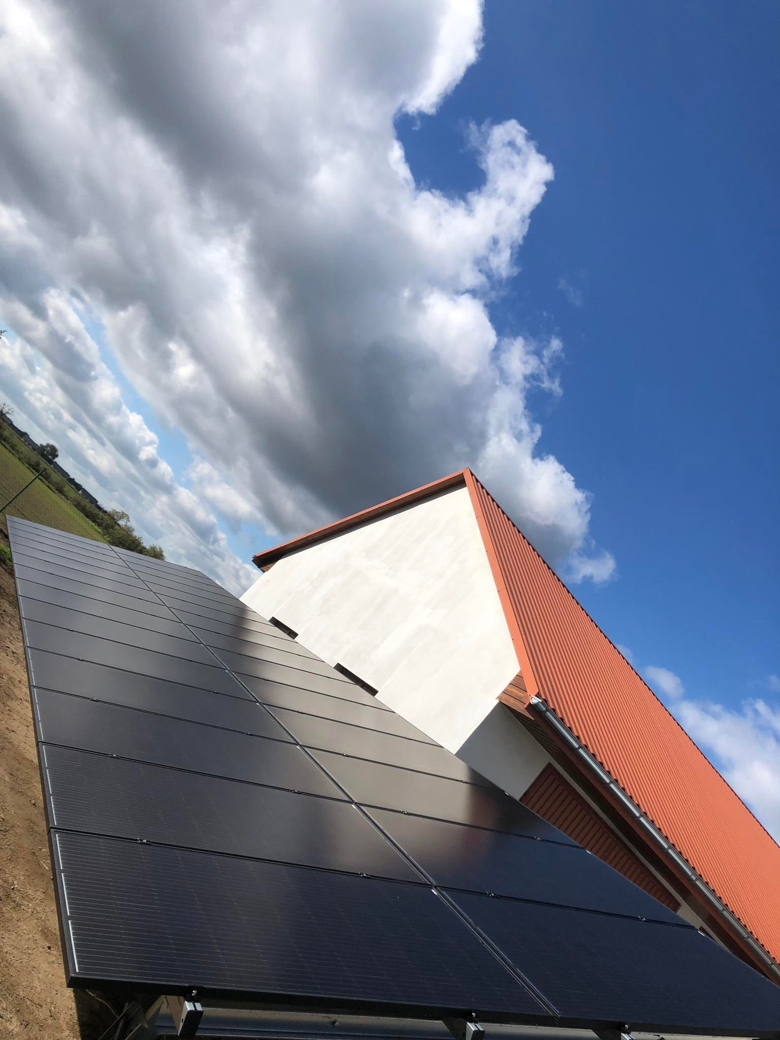 Czarny panel słoneczny zamontowany na gruncie obok białego budynku z czerwoną dachówką, widok pod kątem na tle błękitnego nieba z chmurami.