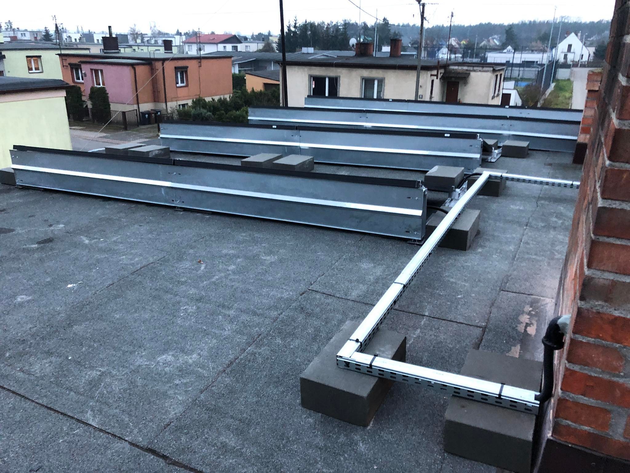 Montaż stelażu pod panele słoneczne na płaskim dachu pokrytym papą, widoczne metalowe profile i betonowe bloczki, w tle zabudowa miejska przy pochmurnej pogodzie.