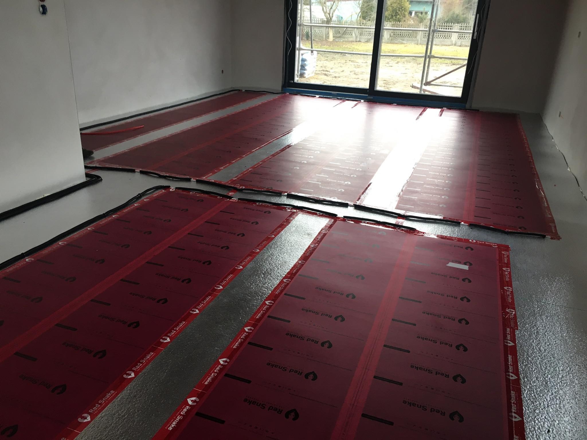 Rozłożone maty grzewcze Red Snake na folii aluminiowej, przygotowane do montażu ogrzewania podłogowego w surowym wnętrzu z dużym oknem.
