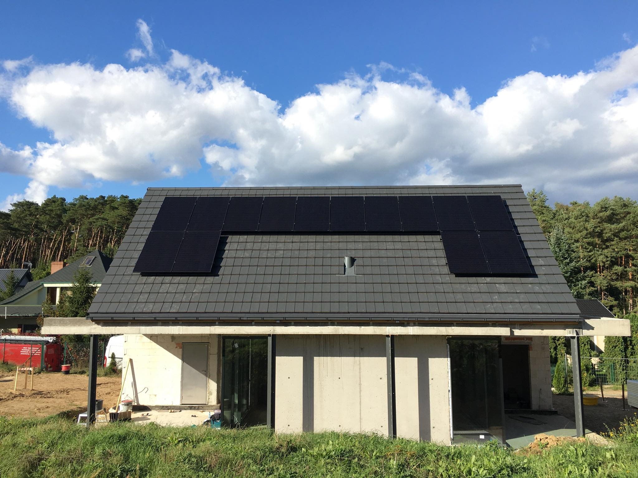 Dom w trakcie budowy z zamontowanymi panelami słonecznymi na szarym dachu z dachówki, błękitne niebo z białymi chmurami w tle.