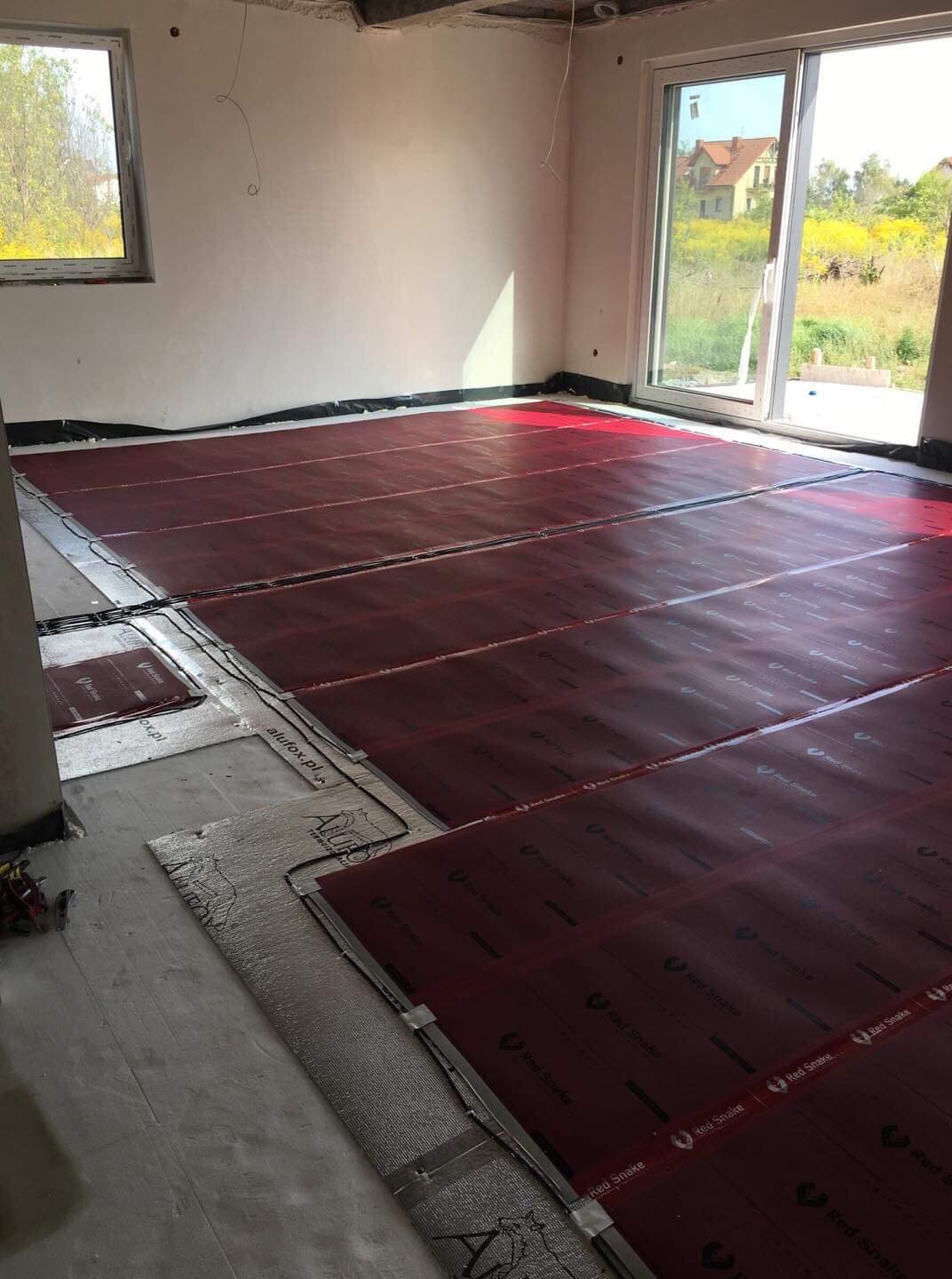 Montaż czerwonych mat grzewczych Red Snake na podłodze w surowym pomieszczeniu z widokiem na ogród przez okno balkonowe.