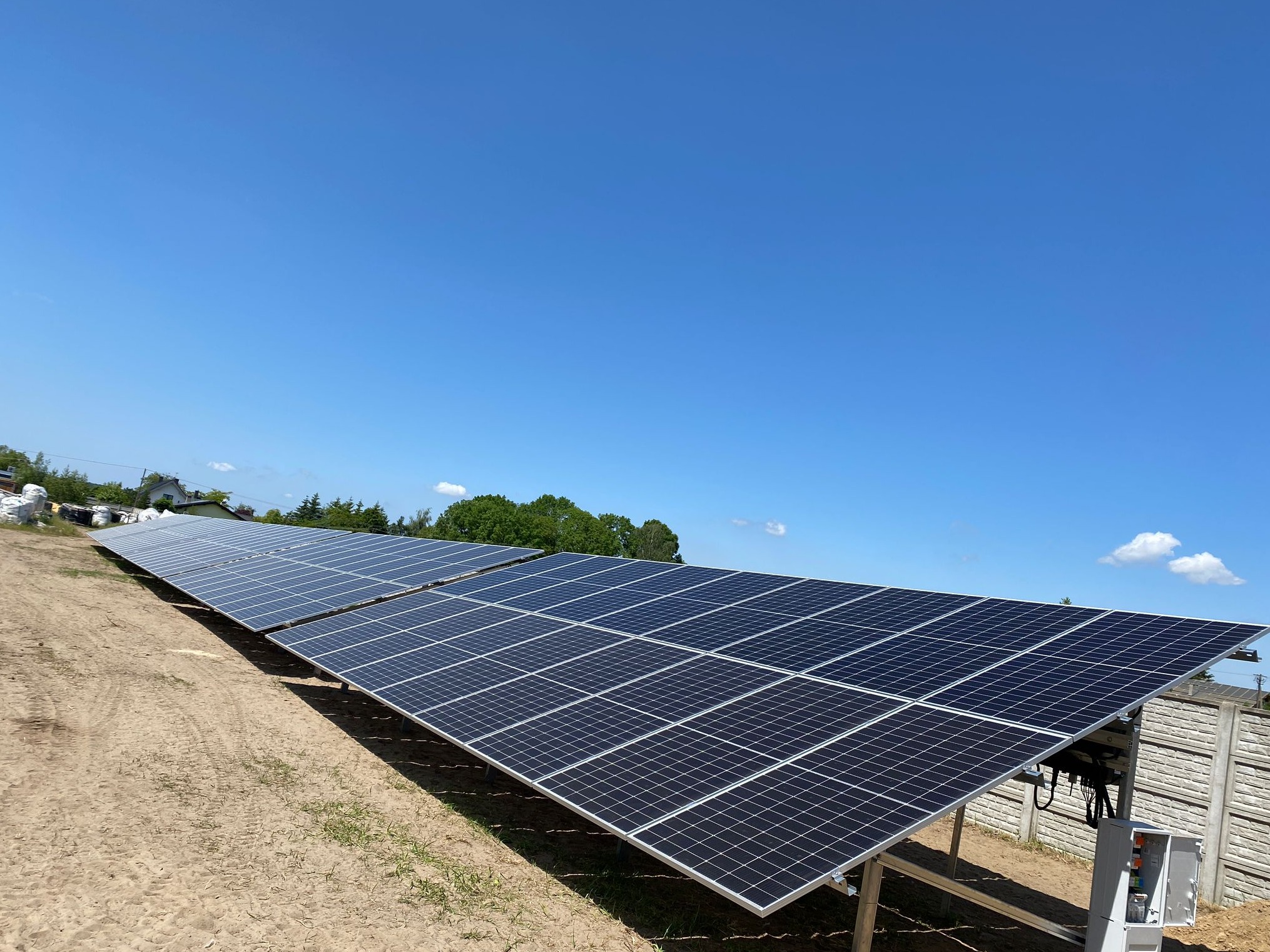 Długa instalacja paneli słonecznych na metalowych stelażach, widoczne kable i skrzynka elektryczna, w tle błękitne niebo i zieleń drzew.