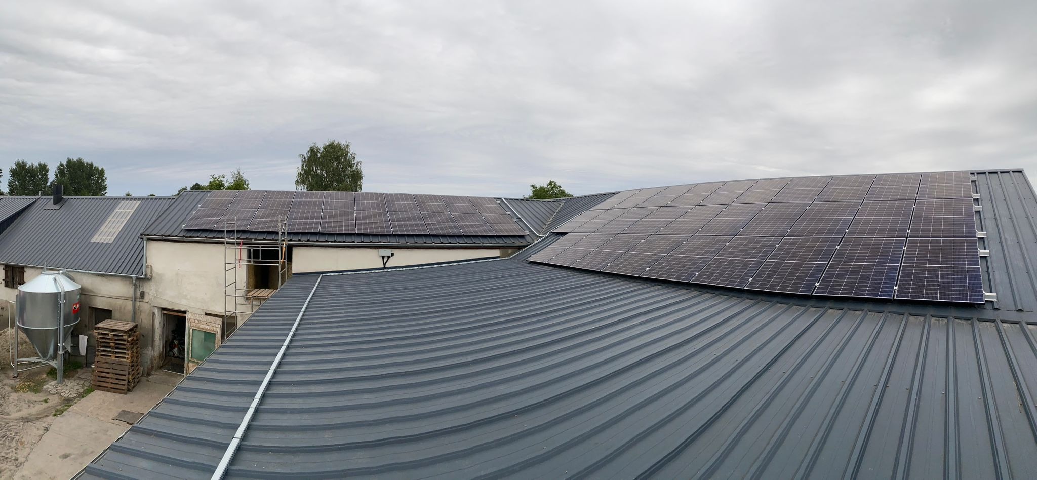 Panele słoneczne zamontowane na falistym, metalowym dachu budynku gospodarczego, widoczne rusztowanie i silos.