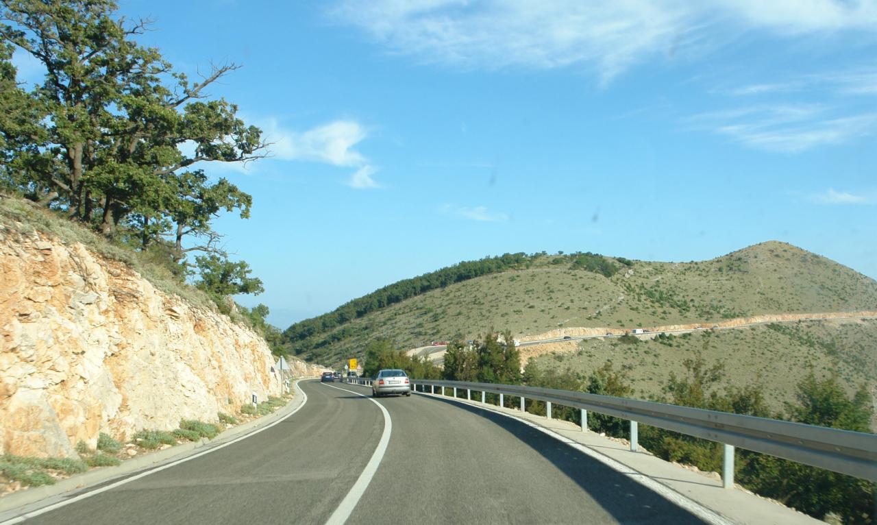 Kręta droga asfaltowa z barierami ochronnymi, prowadząca przez górzysty teren z widocznymi drzewami i krzewami, pod błękitnym niebem z delikatnymi chmurami.