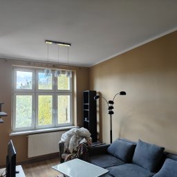Krzysztof Pęcek Firma Budowlana - Jasne wnętrze po malowaniu: beżowe ściany, okno z widokiem na drzewa, szara sofa i nowoczesna lampa. Minimalistyczny design z akcentem na naturalne światło.
