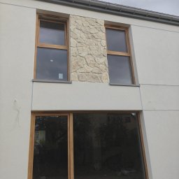 Krzysztof Pęcek Firma Budowlana - Fragment elewacji budynku z oknami w drewnianych ramach i dekoracyjnym panelem z kamienia. Świeżo otynkowana ściana w jasnym odcieniu. Widoczne ślady prac wykończeniowych.