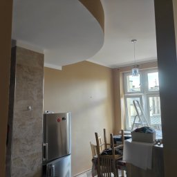 Krzysztof Pęcek Firma Budowlana - Beżowe ściany po malowaniu w mieszkaniu z zaokrąglonym sufitem, widoczne meble i drabina przy oknie. Ściana wykończona płytkami imitującymi marmur.