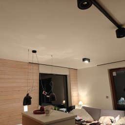 Krzysztof Pęcek Firma Budowlana - Jasne wnętrze pokoju z drewnianą ścianą, lampami wiszącymi i szynoprzewodem oświetleniowym. Widoczny fragment sofy i drzwi balkonowe.
