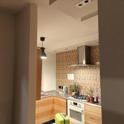 Krzysztof Pęcek Firma Budowlana - Jasna kuchnia z drewnianymi szafkami, mozaiką na ścianie i nowoczesnym okapem. Na blacie rośliny w doniczkach. W tle widoczny fragment salonu z lampą.