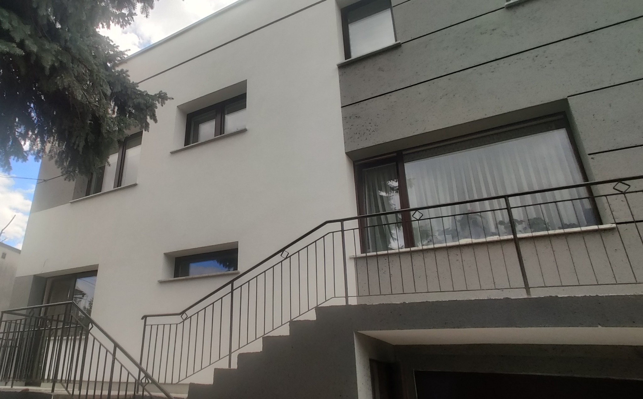 Dwukondygnacyjny budynek z elewacją w kolorze białym i szarym, z zewnętrznymi schodami z metalową balustradą i widocznym drzewem iglastym po lewej stronie.