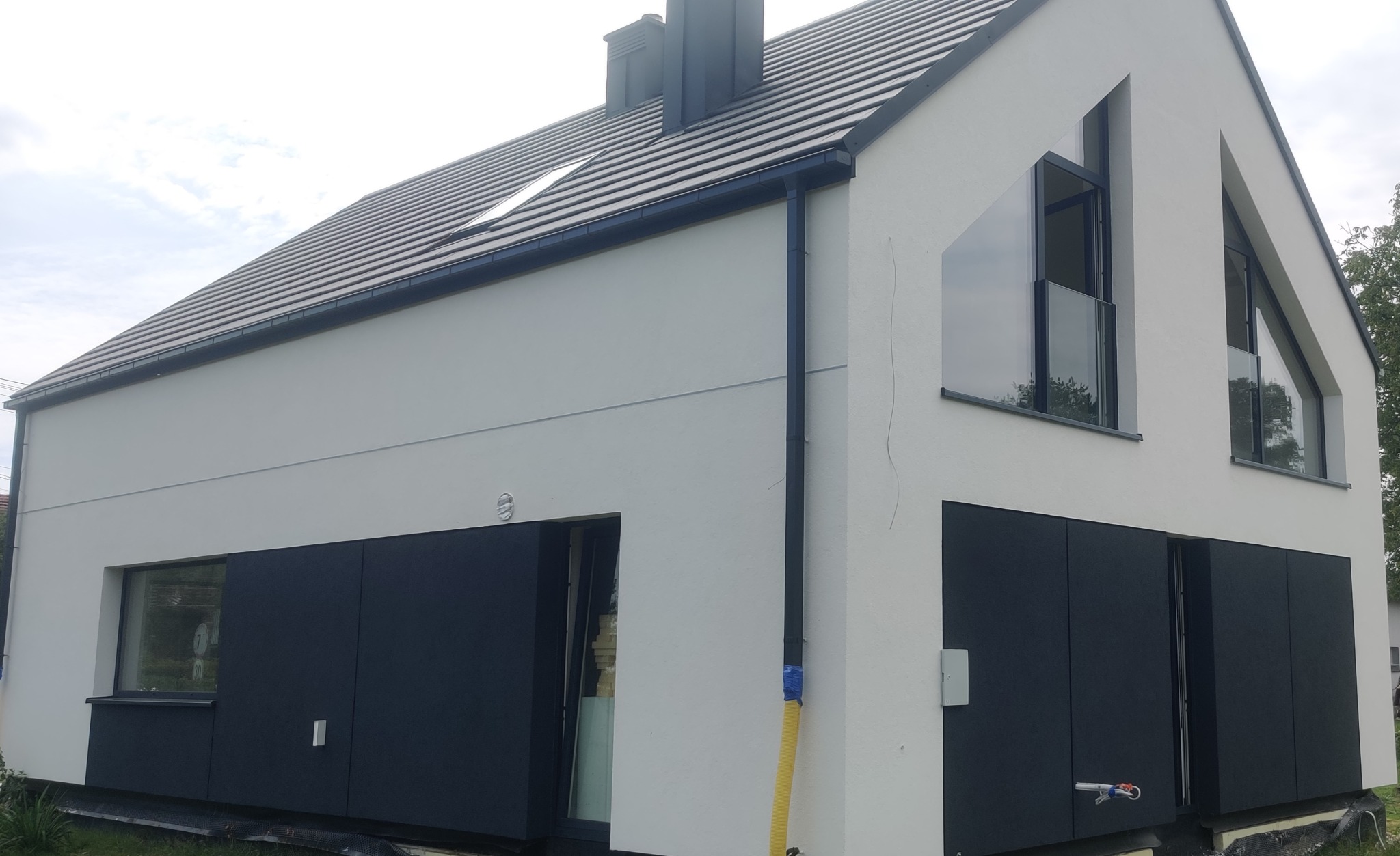 Nowoczesny dom jednorodzinny z białą elewacją, antracytowymi panelami dolnymi, oknami i dachem, z widoczną rurą gazową w żółtej osłonie.