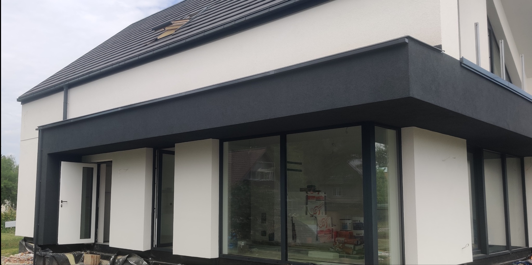 Nowoczesny dom z białą elewacją i grafitowym wykończeniem, widoczne okna i otwarte drzwi, teren wokół budynku w trakcie prac wykończeniowych.