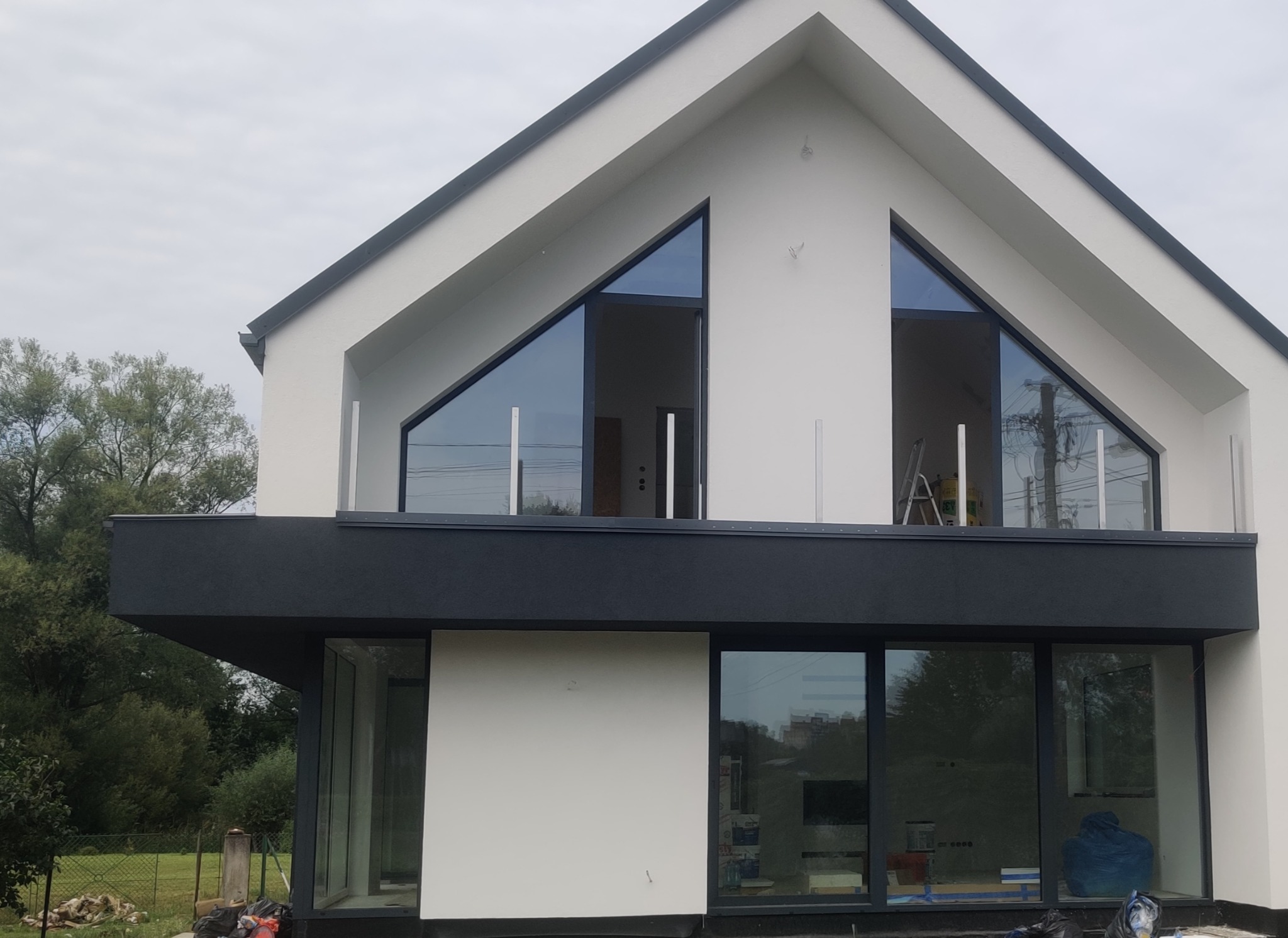 Nowoczesny dom z białą elewacją i czarnymi akcentami, widoczne duże okna trójkątne i balkon z szklaną balustradą, na balkonie stoi drabina.